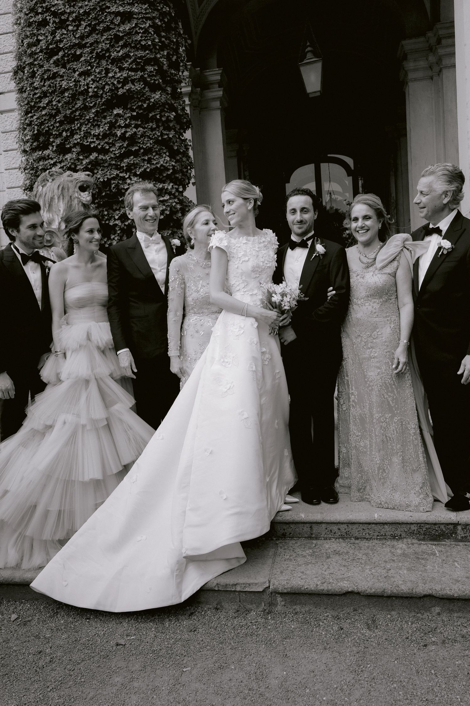 Black and white wedding photo of bride in elegant gown with groom and family on steps of ivy-covered estate.