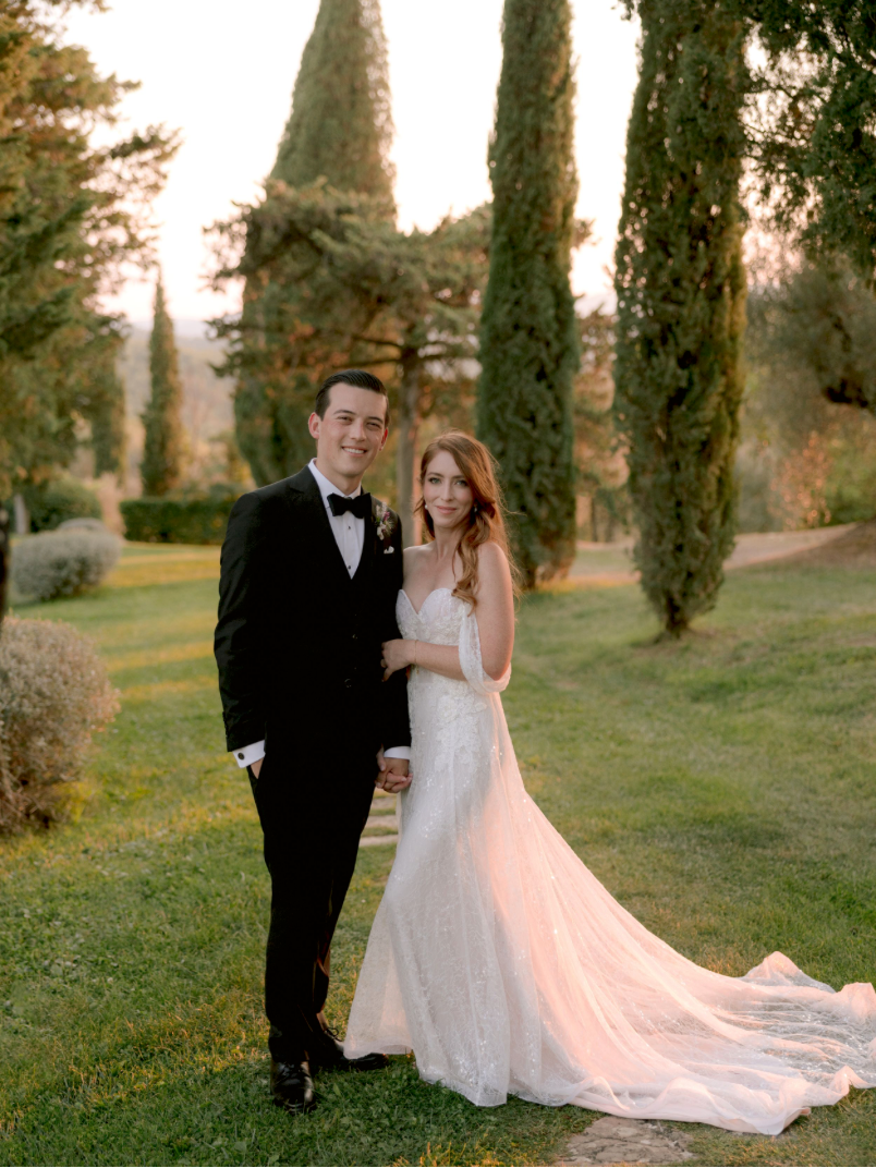 Bride and groom pose together in a sunlit garden with tall cypress trees at their elegant outdoor wedding.