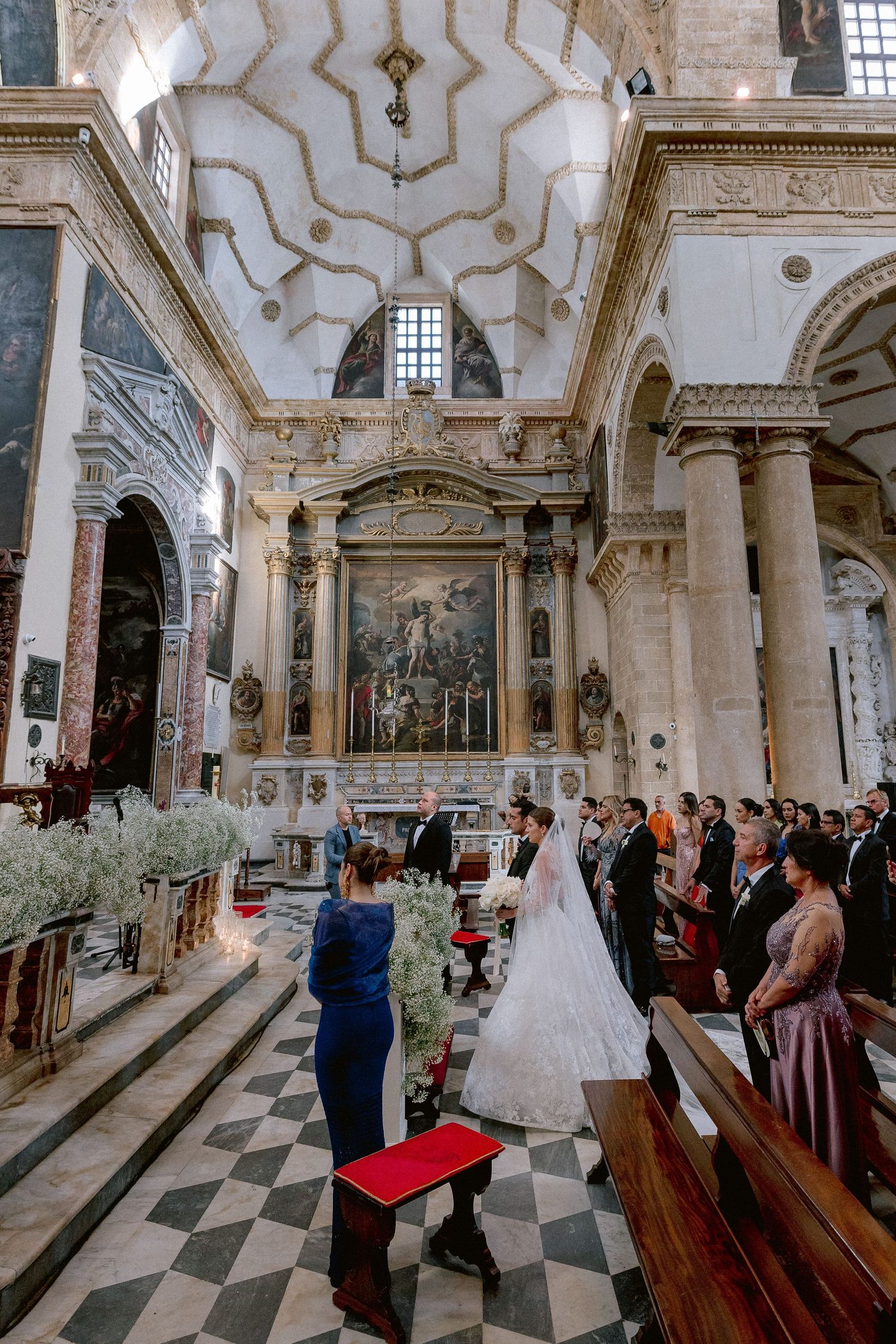 Ornate Italian baroque church interior with checkered floor and floral decorations during a wedding ceremony.
