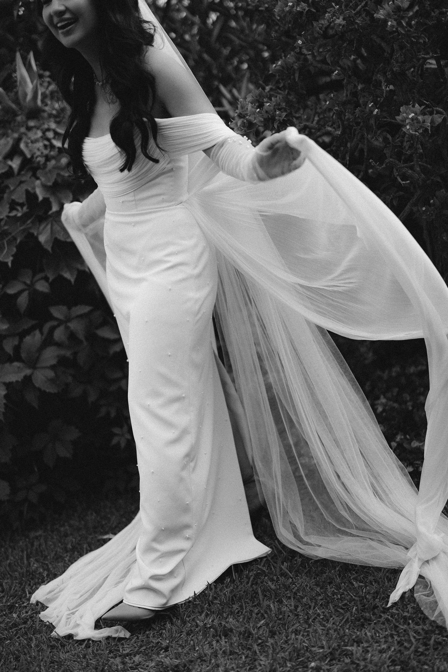 Black and white photo of an elegant wedding dress with flowing tulle train billowing in garden setting.