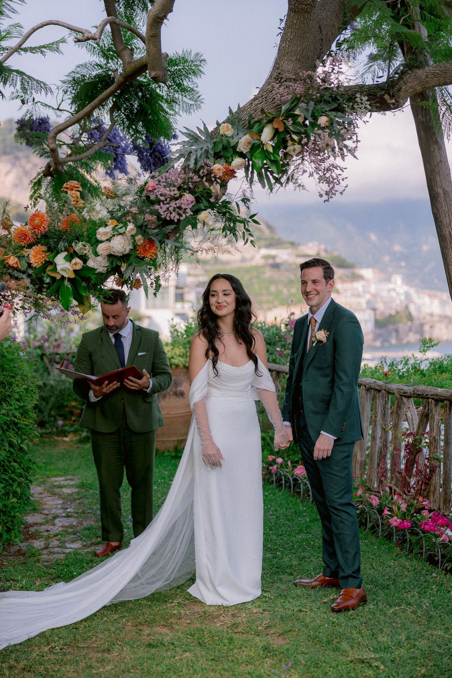 A wedding ceremony takes place on a garden terrace overlooking a coastal town with floral arch and white decor.
