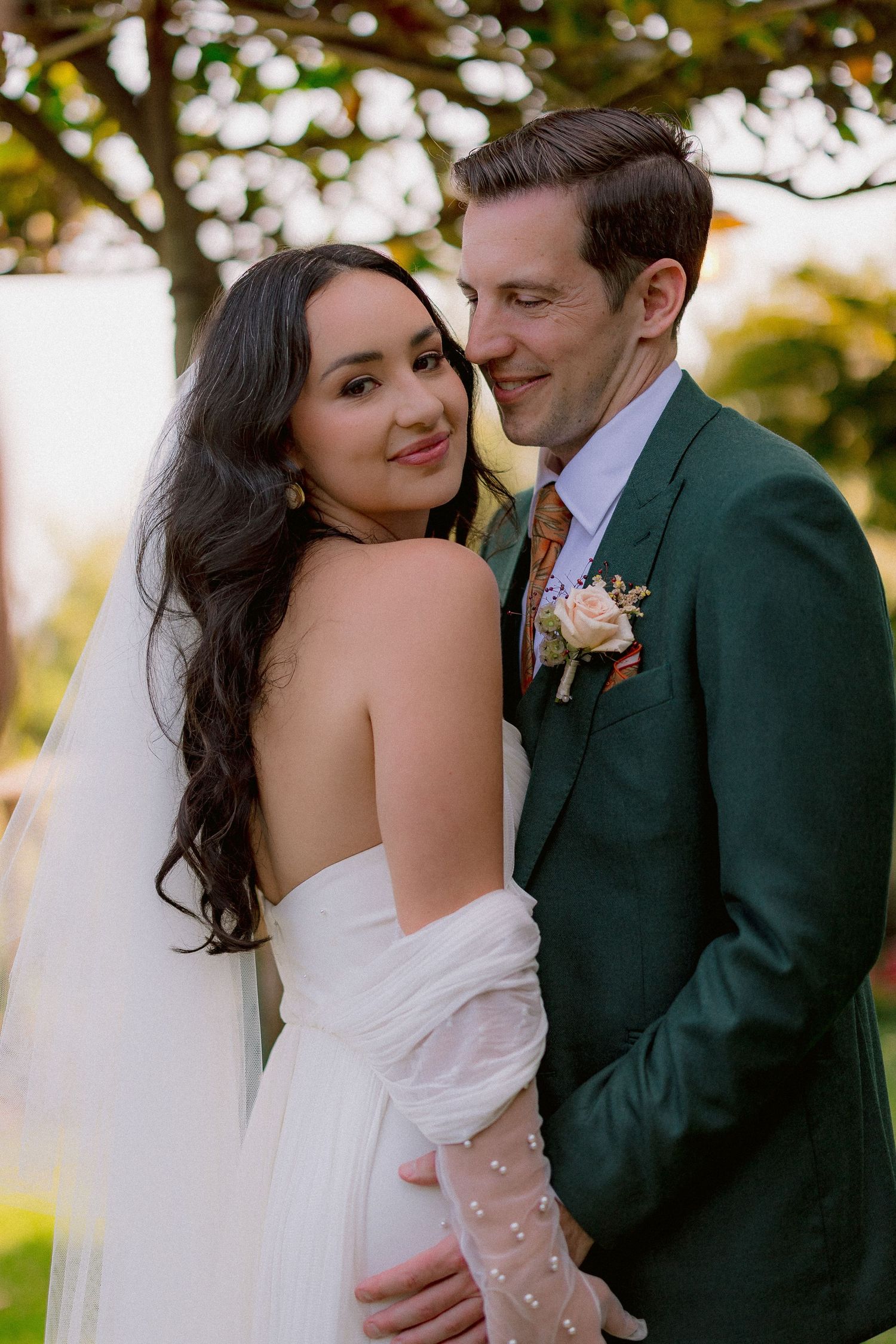 A romantic wedding couple embraces outdoors, with the bride in white and groom in green suit with floral boutonniere.