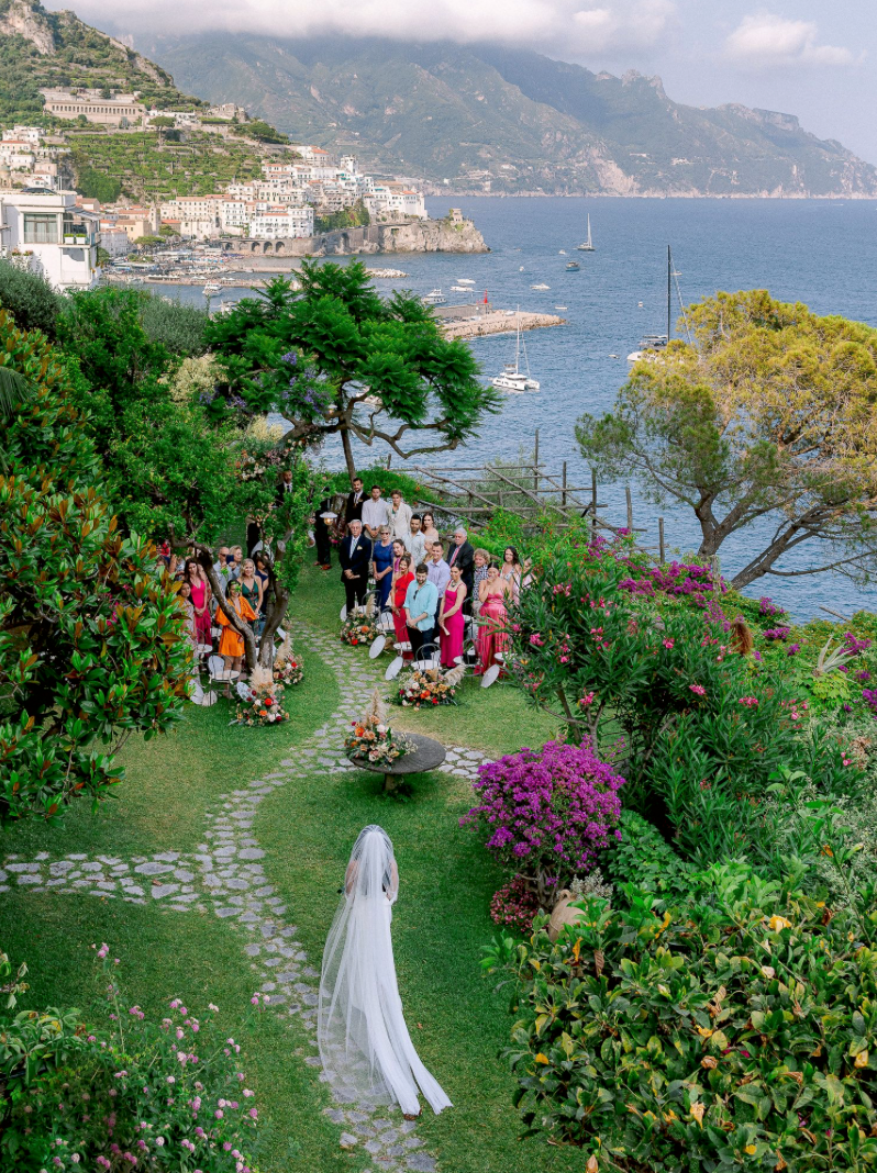 Bride in white gown and veil walks toward wedding guests in lush garden overlooking Amalfi Coast, Italy.