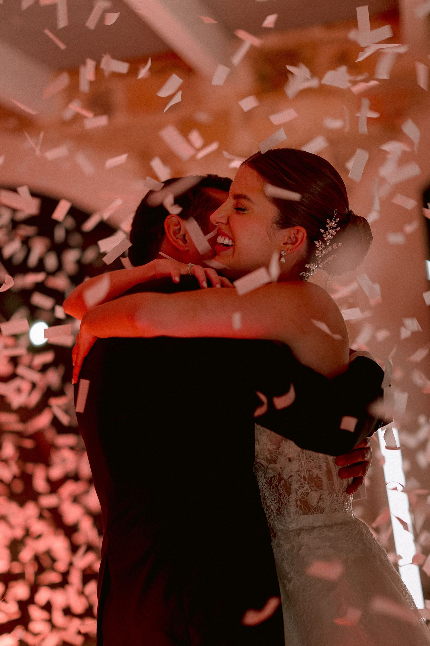A couple embraces during their first dance at a wedding reception as confetti falls around them in warm lighting.