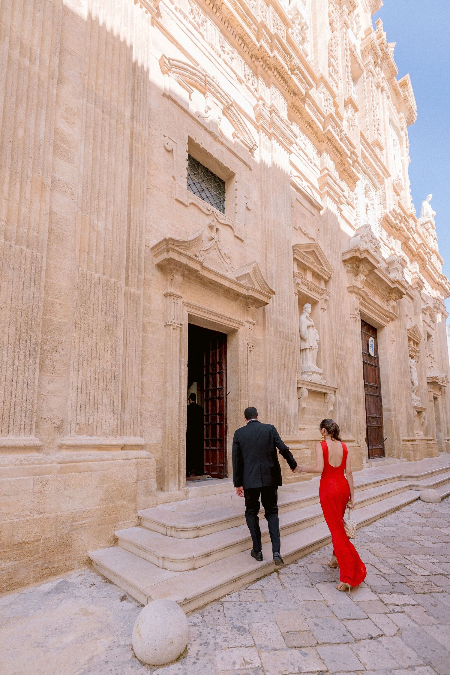 Two people walking up ornate limestone steps of a historic baroque church in Gallipoli in formal attire.