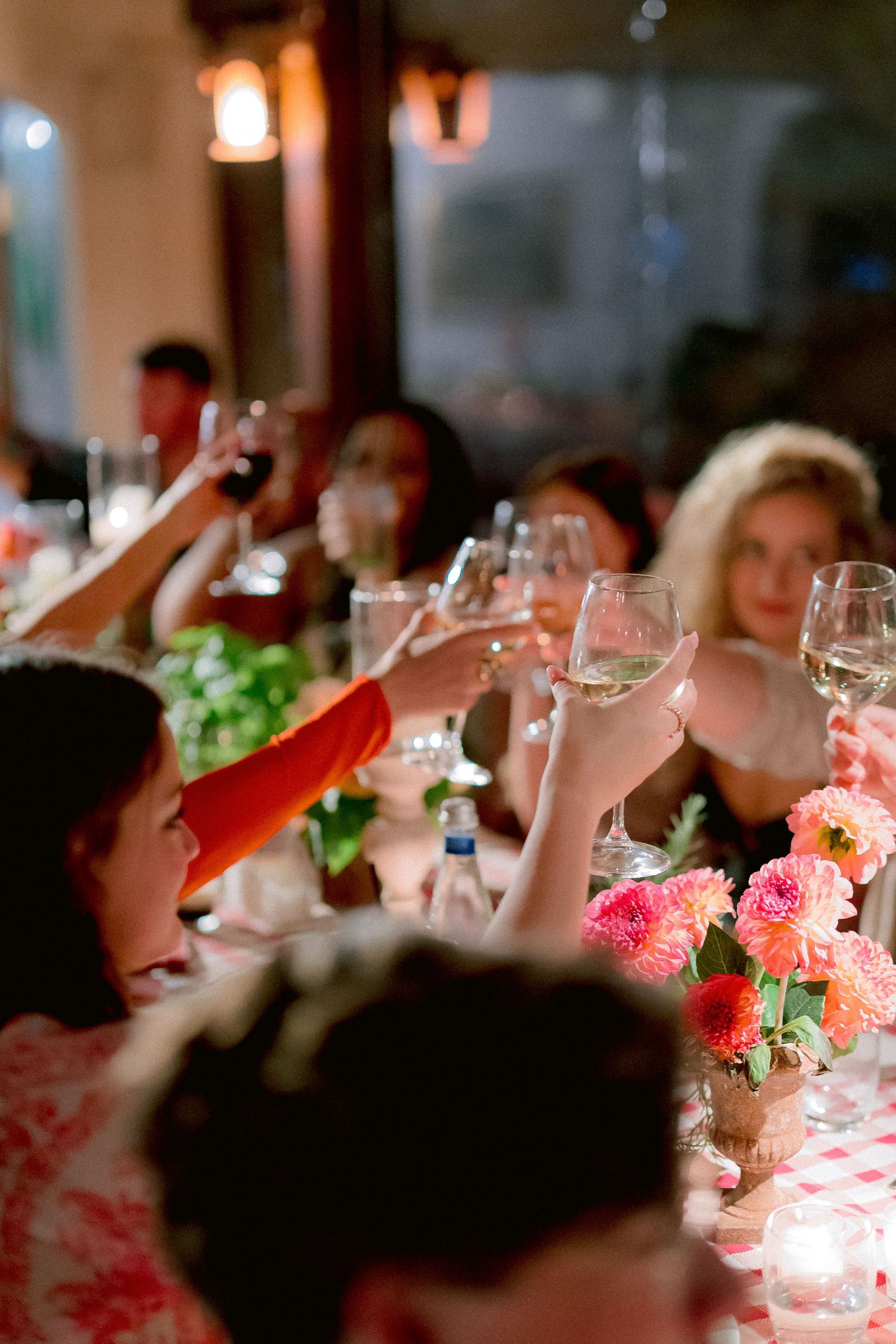 People raising wine glasses in a toast at a dinner party with pink floral centerpieces and warm ambient lighting.