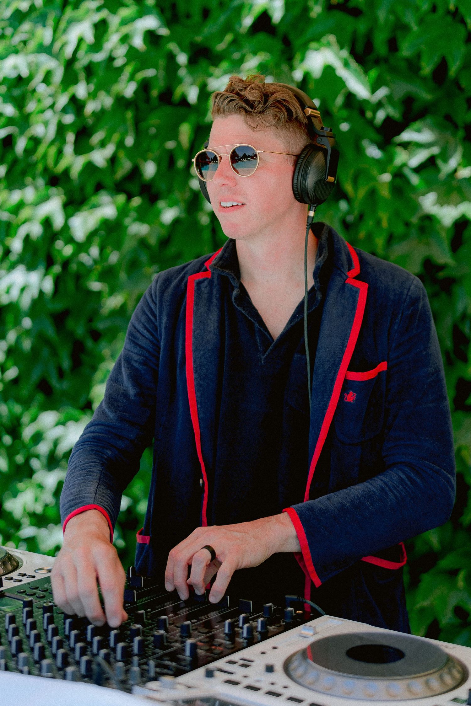 DJ wearing sunglasses and navy blazer with red trim performs at outdoor event with turntables and green foliage backdrop.