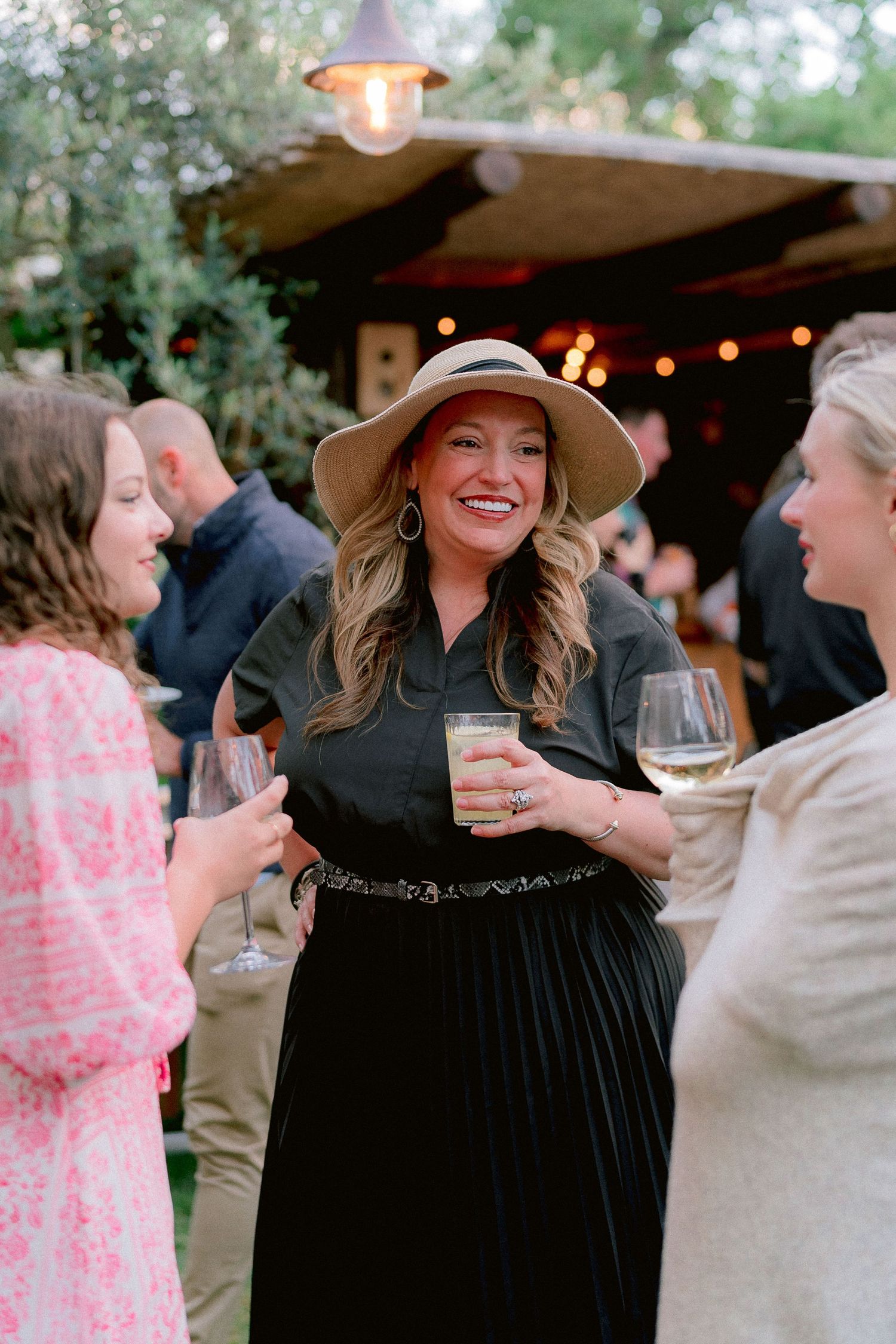 At an outdoor party with string lights, people socialize while holding drinks under a covered patio area.