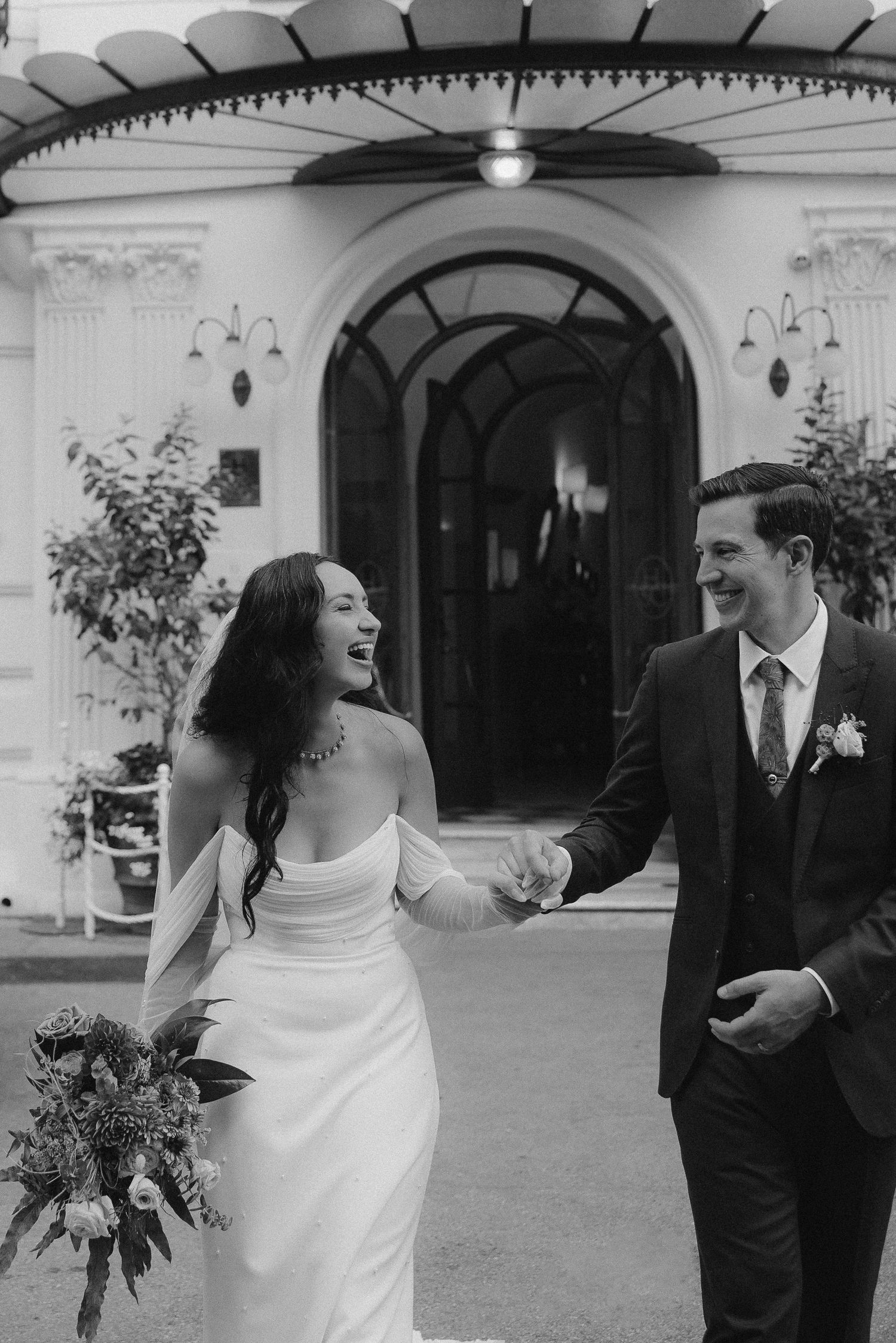 A sweet black and white wedding photo shows a happy couple laughing together outside a beautiful arched entryway.