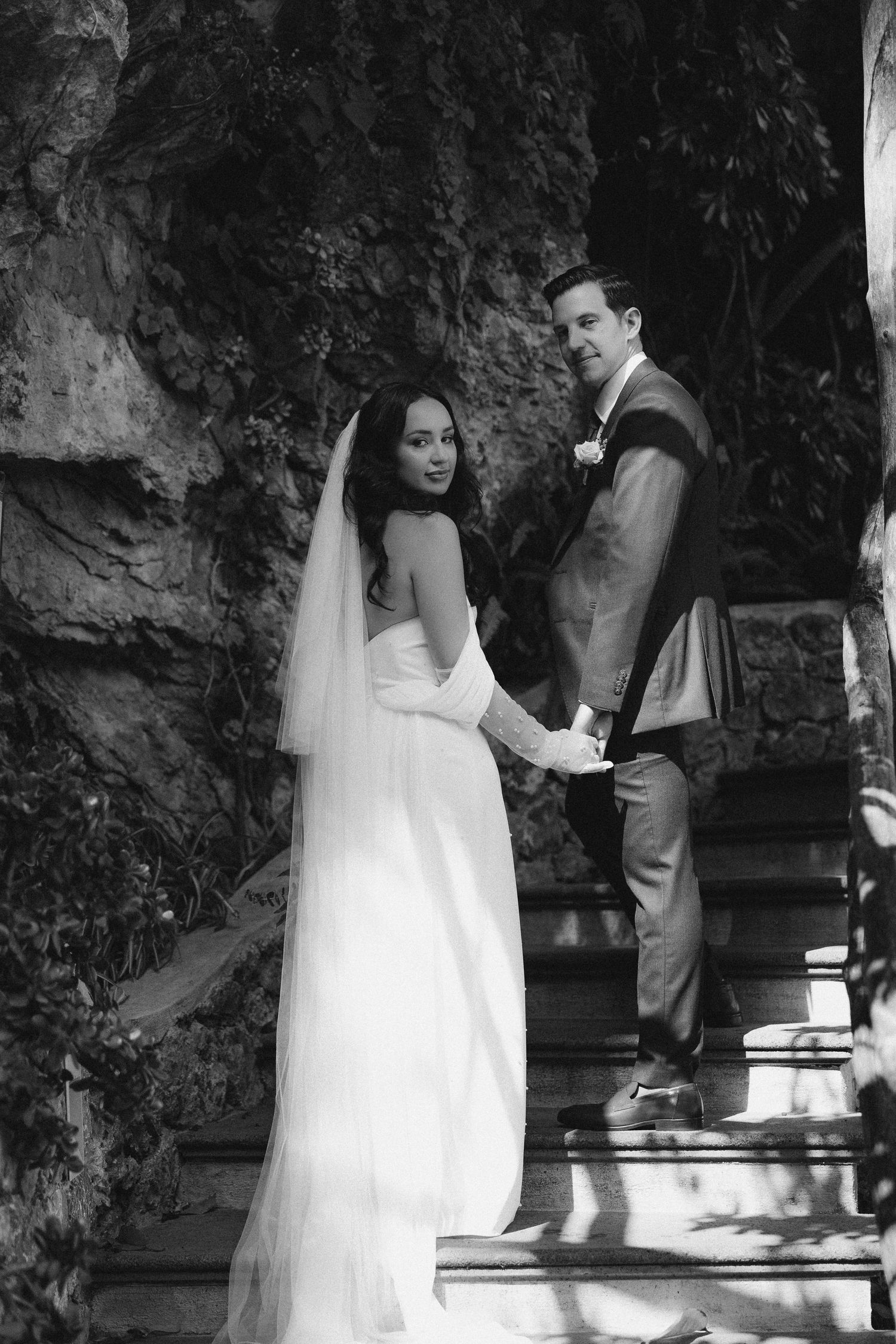 Bride and groom pose on outdoor stone steps in elegant black and white wedding photo.
