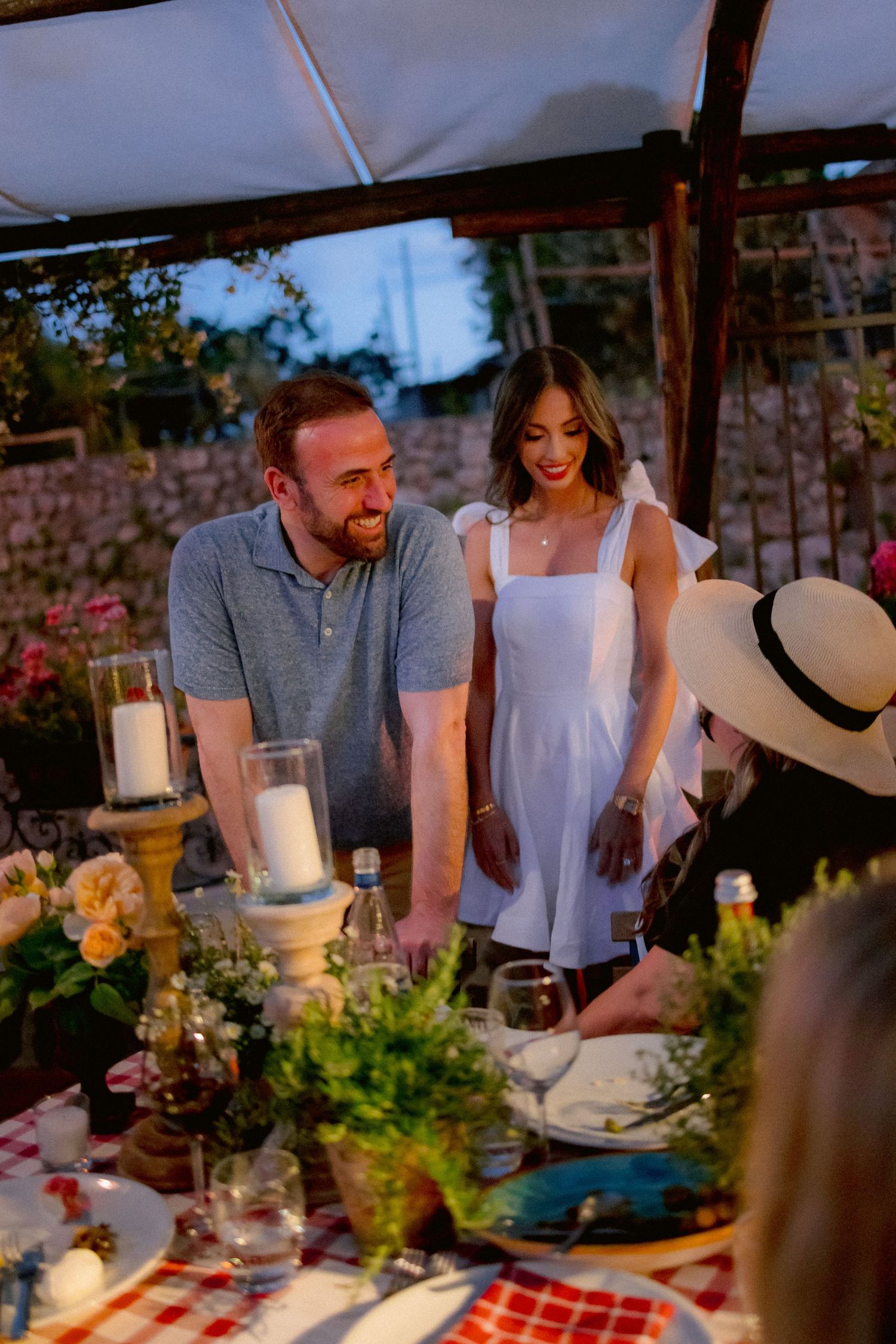 A couple stands at an outdoor dining table set with candles and flowers during a warm evening gathering.
