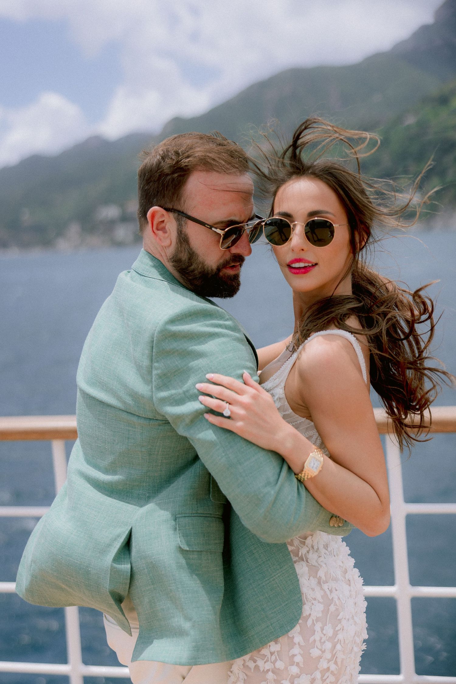 A romantic couple embraces on a cruise ship deck with scenic mountains and water in the background.