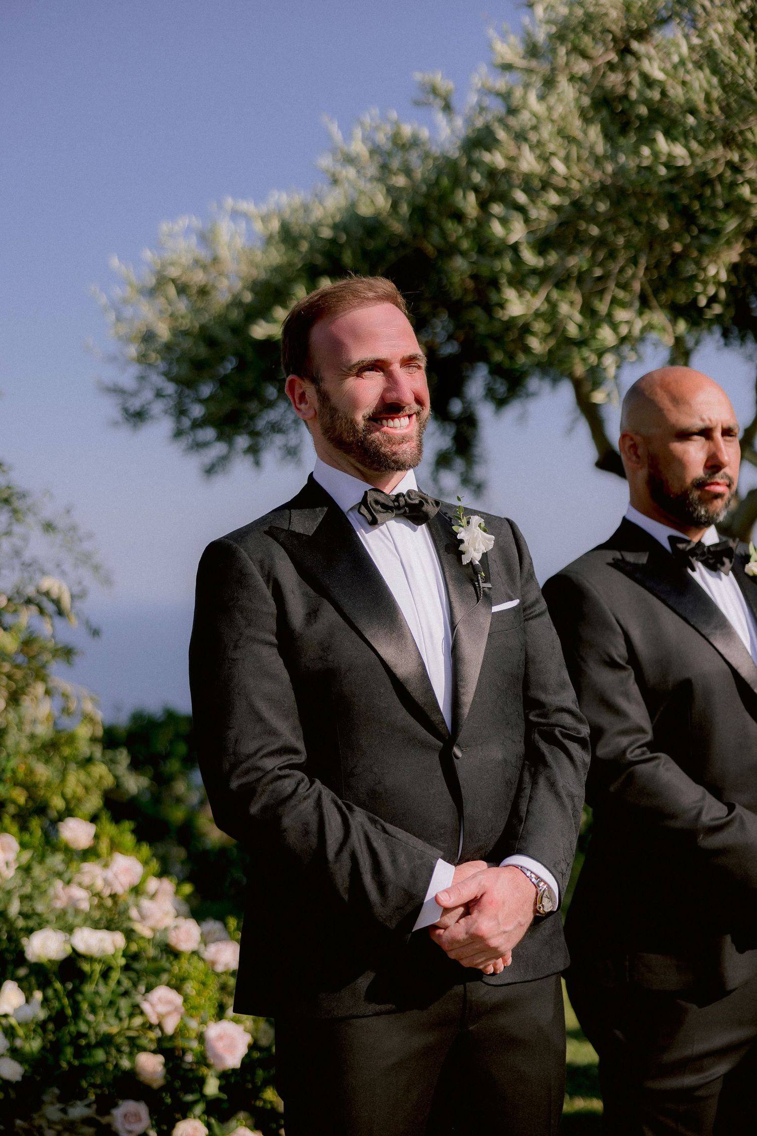 Two groomsmen in classic black tuxedos and bow ties stand at an outdoor wedding ceremony with flowering shrubs behind them.