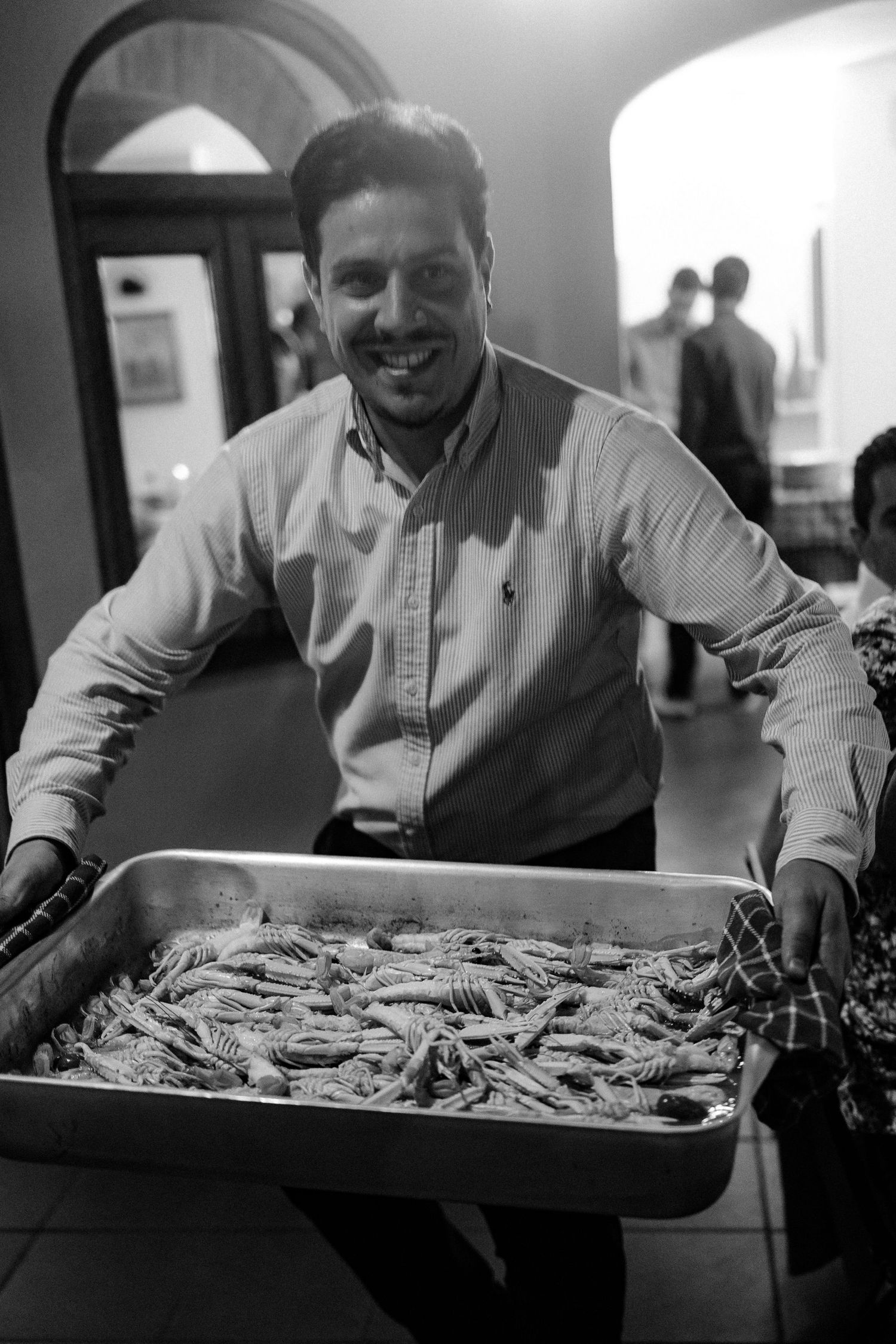 A person in a button-down shirt smiles while holding a large tray of fresh seafood in a black and white photo.