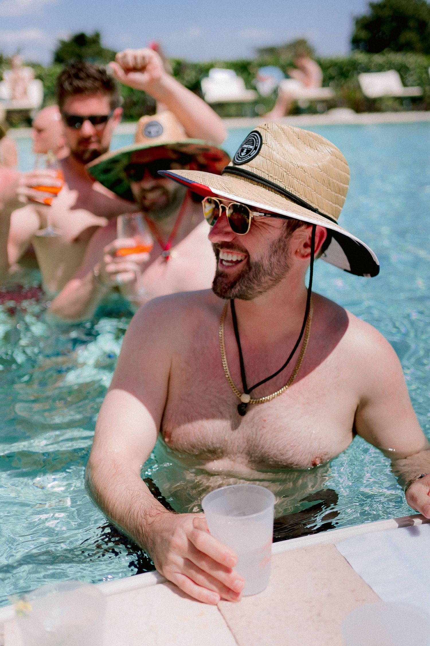 People enjoying drinks and socializing in a swimming pool on a sunny day while wearing sunglasses and a straw hat.