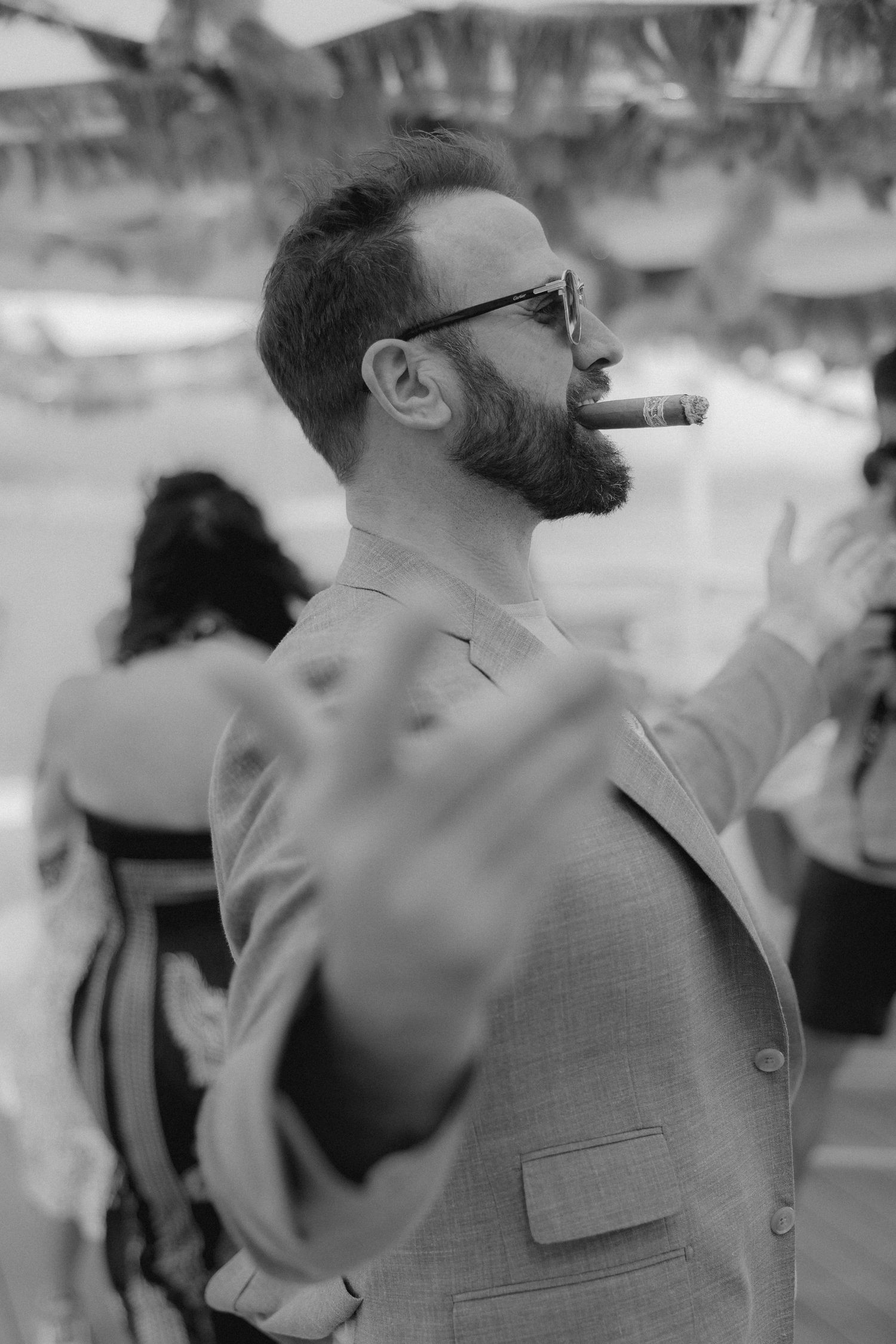 Black and white vintage style photo of a bearded man in a suit jacket smoking a cigar at an outdoor event.