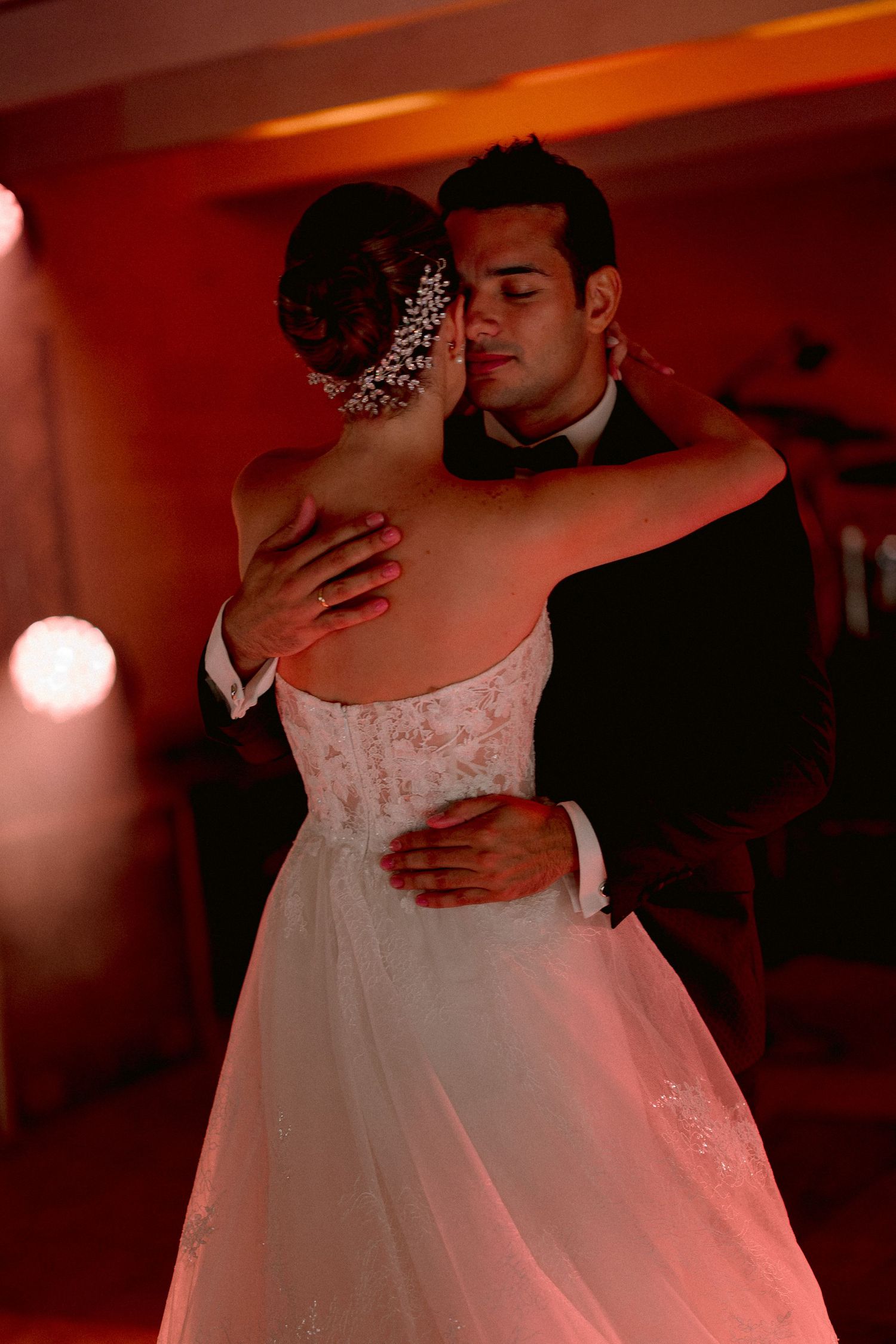 A newlywed couple shares their first dance in warm reddish lighting at their wedding reception.