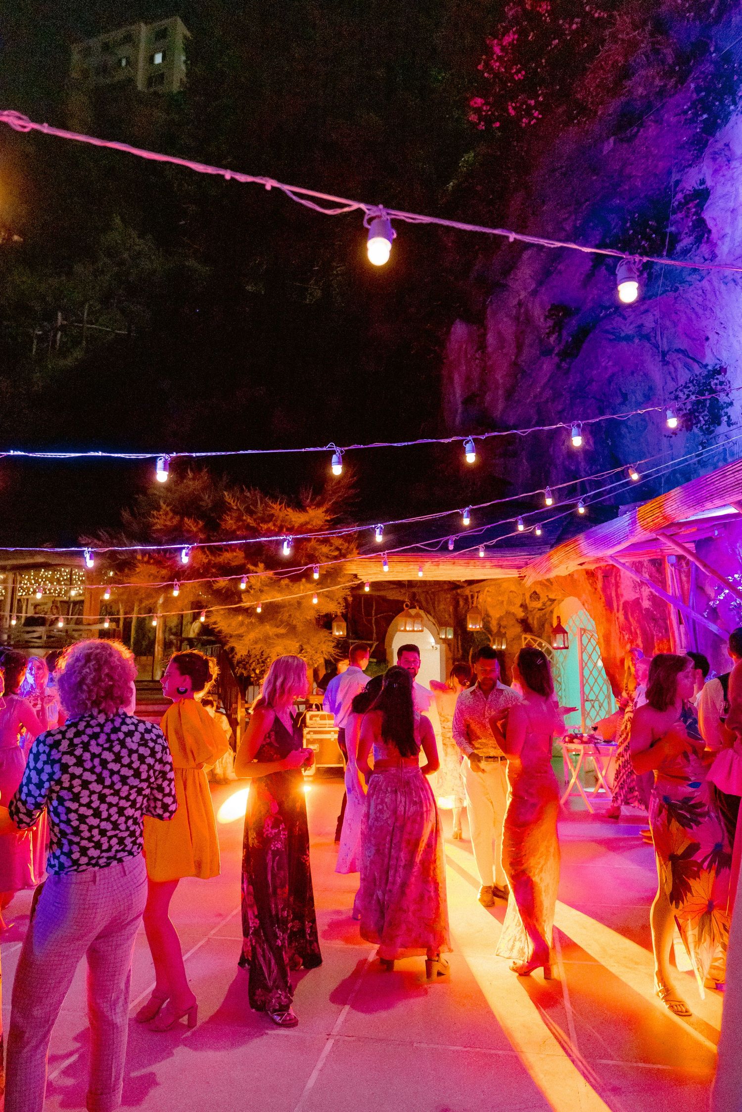 People dancing at an outdoor party illuminated by colorful string lights and purple ambient lighting creating a festive atmosphere.