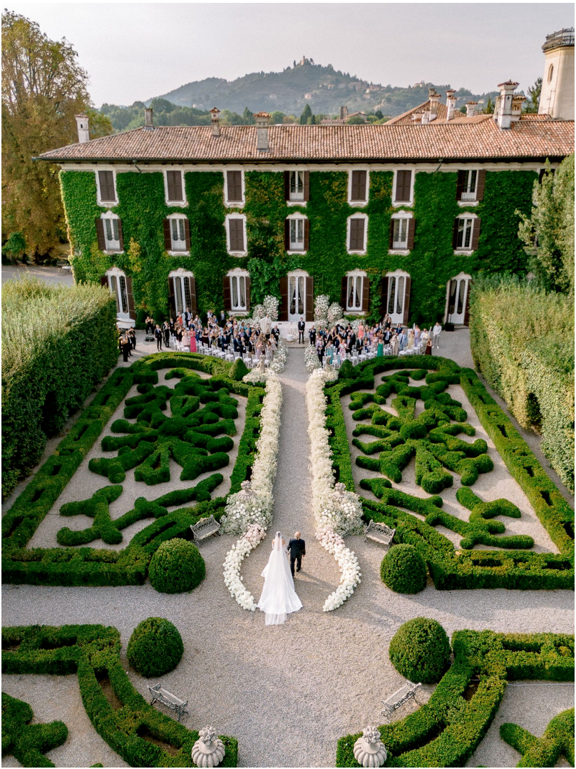 Aerial view of elegant ivy-covered villa wedding ceremony in formal Italian garden with manicured hedges and floral aisle.