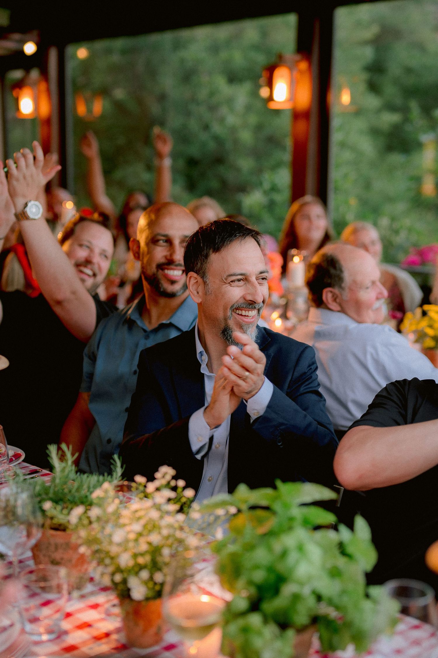 Wedding reception guests laugh and clap during celebration with warm lighting and floral centerpieces on tables.