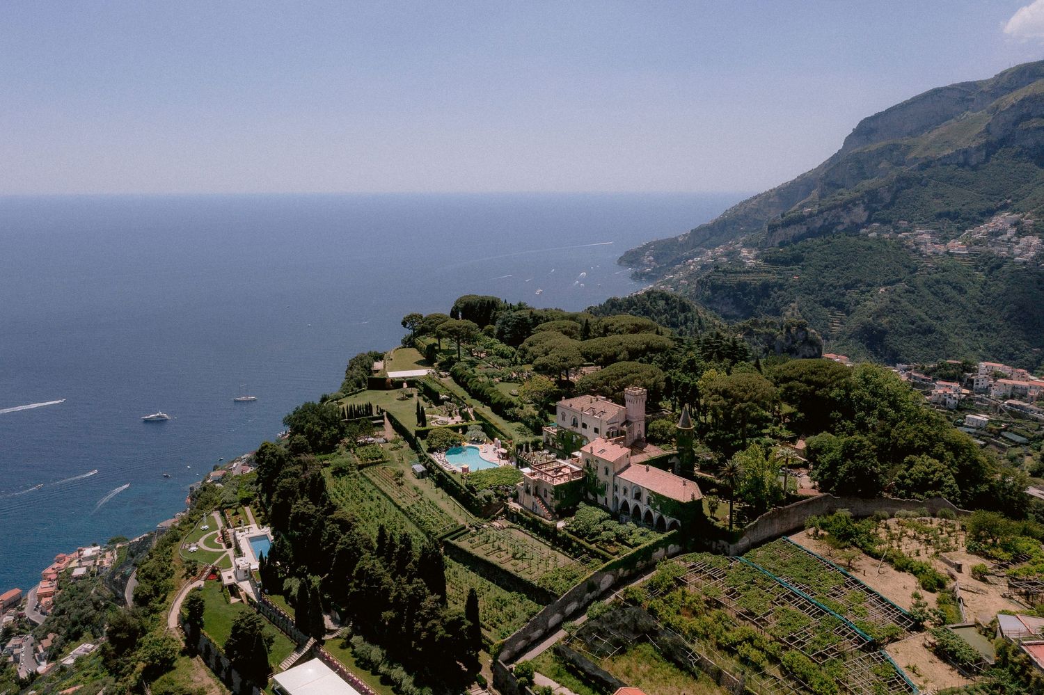 Aerial view of terraced gardens and buildings along the Mediterranean coastline with mountains and blue sea in Sicily.