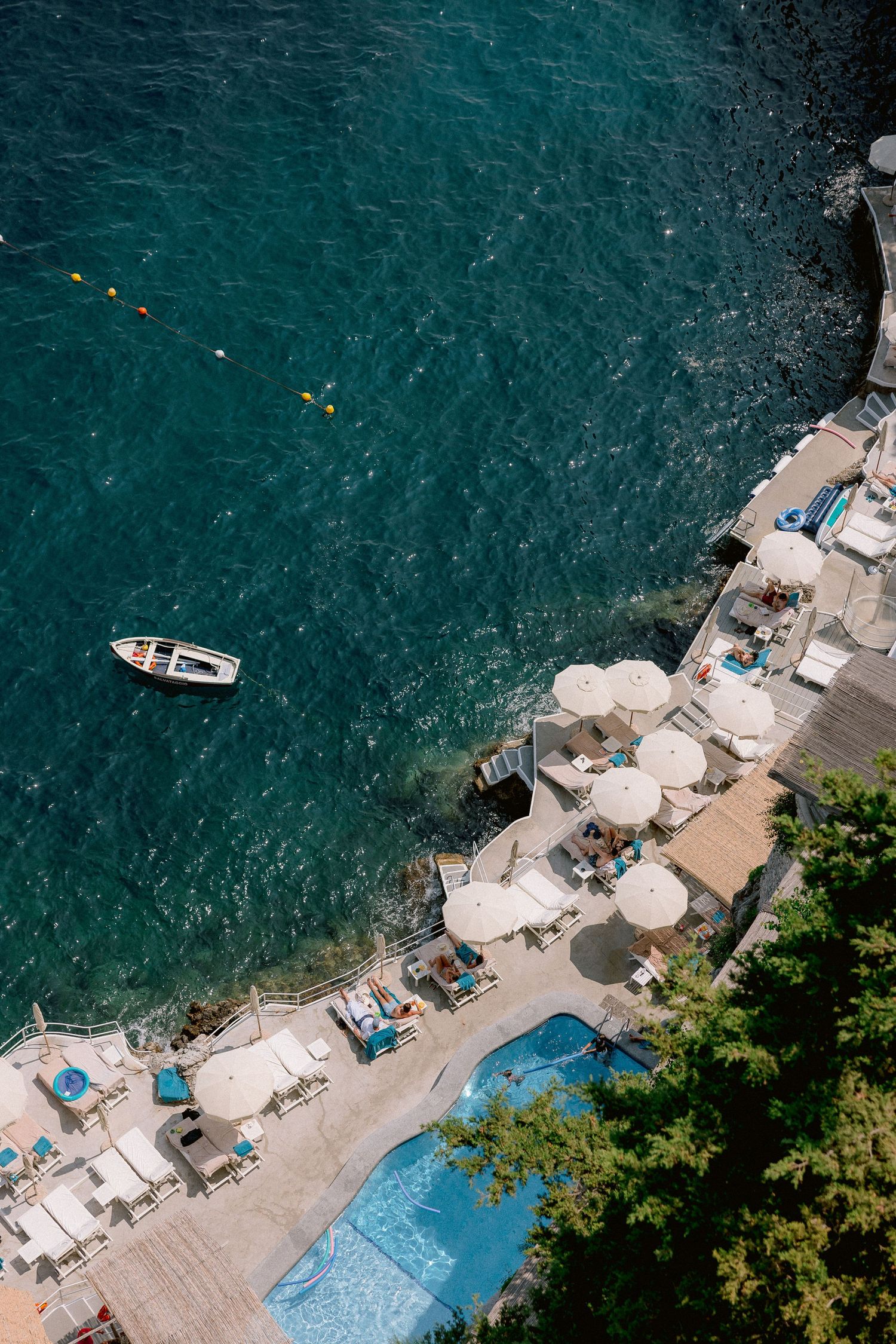 Aerial view of a coastal beach club with turquoise water, small boat, swimming pool and lounge chairs along rocky shoreline.