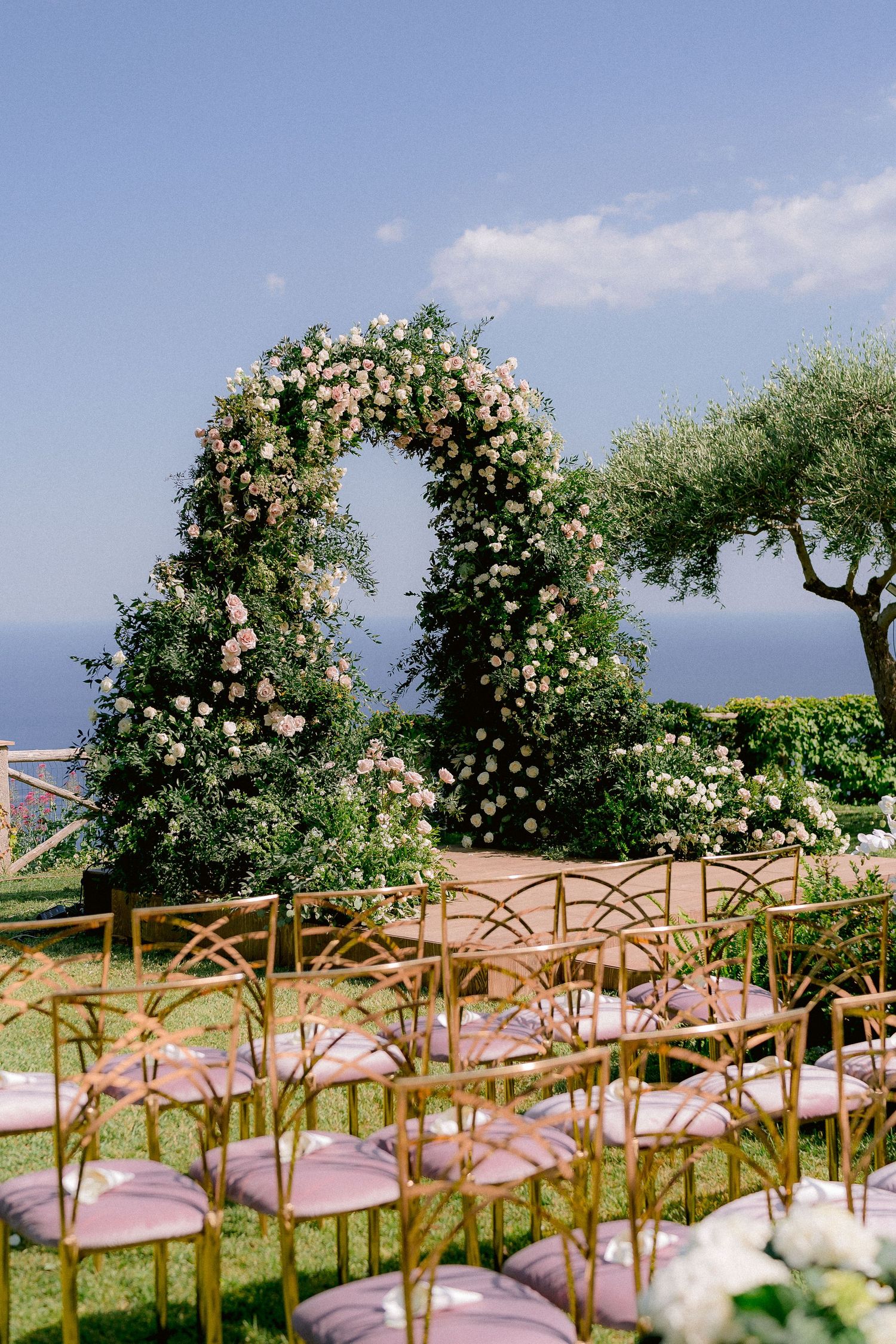 A floral wedding arch with roses overlooks the ocean, with wooden chairs set up for an outdoor ceremony in the garden of the two fountains.