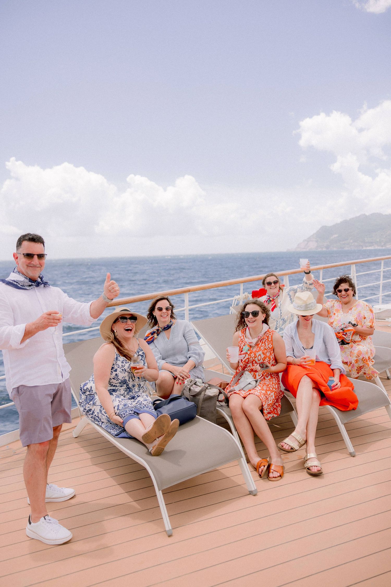 Group of friends enjoying drinks and leisure time together on a cruise ship deck with ocean and mountains in background.