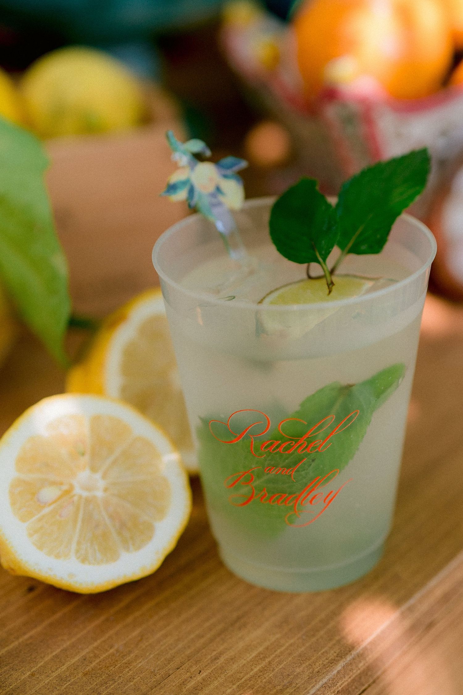 A refreshing frosted cocktail glass garnished with fresh lemon and mint leaves sits beside a sliced citrus on wooden surface.
