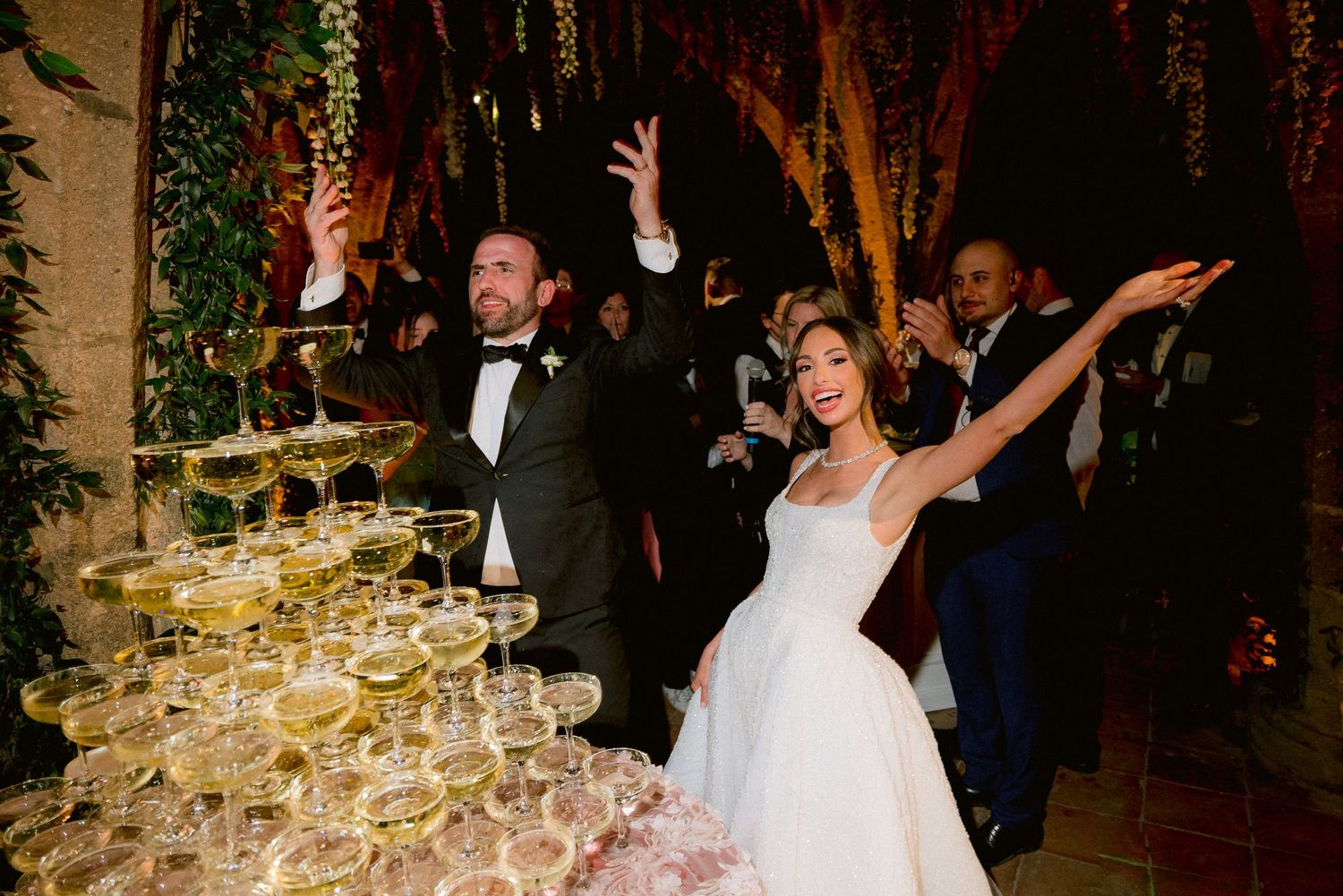 A celebratory moment at a wedding reception with champagne tower and guests raising their arms in joy.