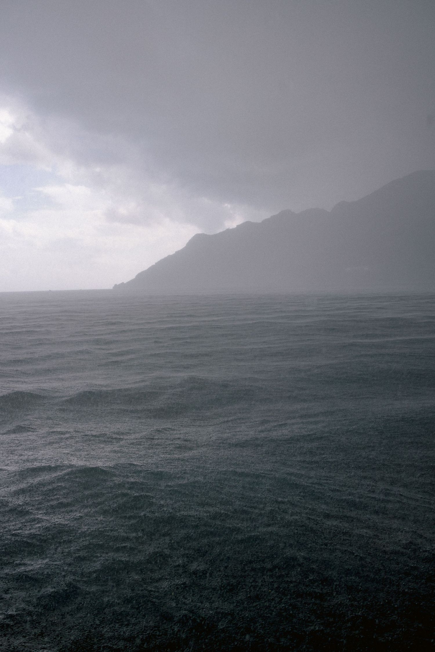 Moody seascape with dark stormy waters against misty mountain silhouettes under overcast skies.