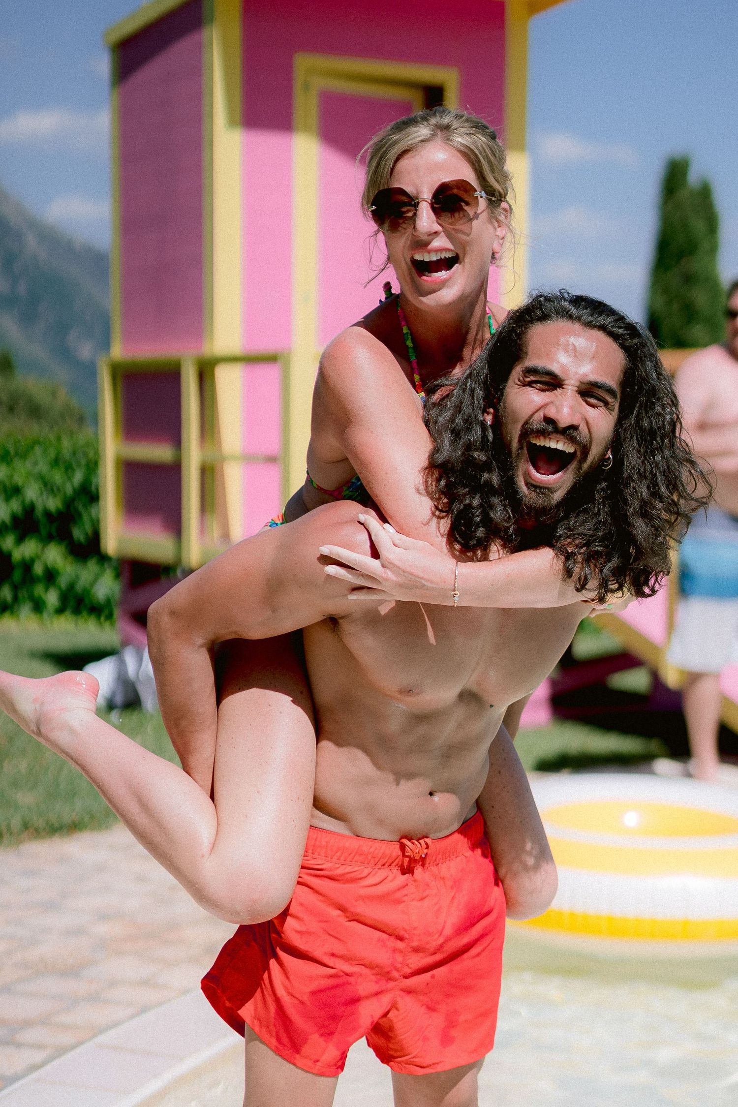 A playful couple shares a piggyback ride moment near a bright pink and yellow lifeguard tower on a sunny summer day.