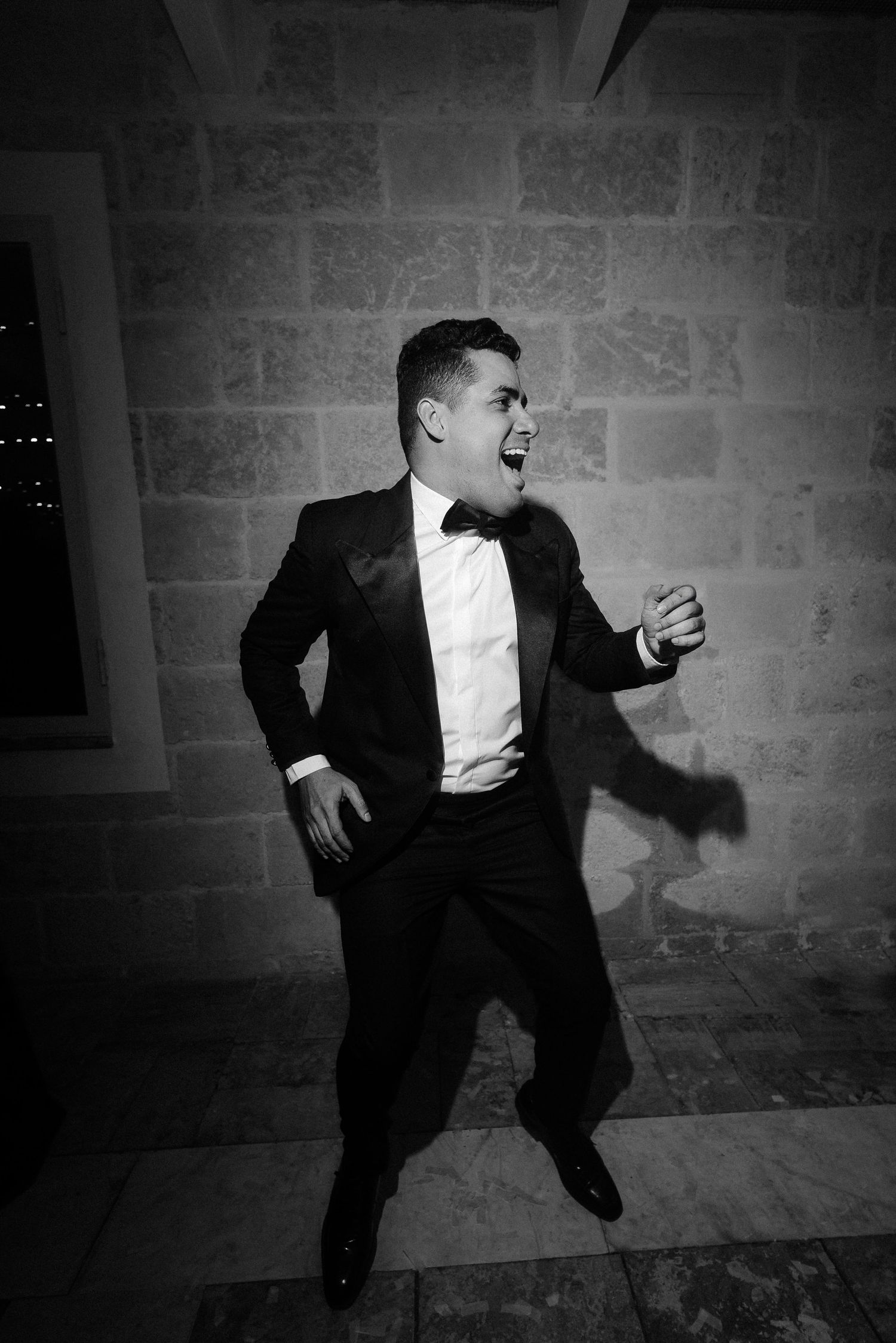 A black and white photo of someone dancing energetically in a tuxedo against a stone wall.