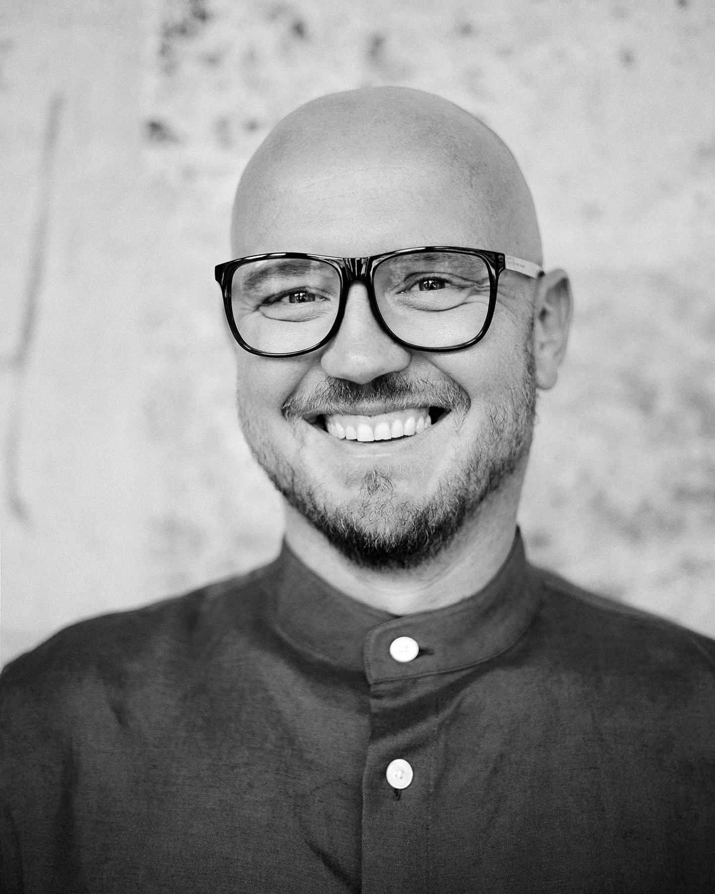 A black and white portrait of a person with glasses, a shaved head and a neatly trimmed beard smiling warmly.