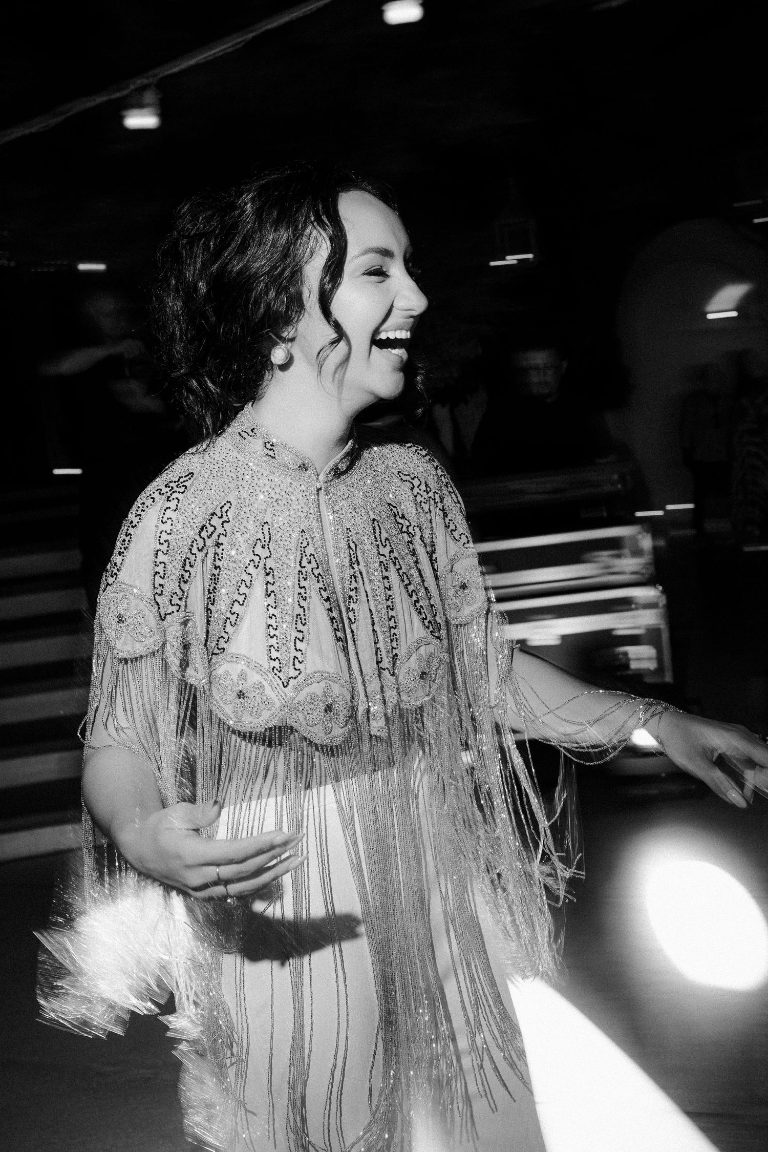 A person in a glamorous fringed dress laughs joyfully at a dimly lit venue in black and white photography.