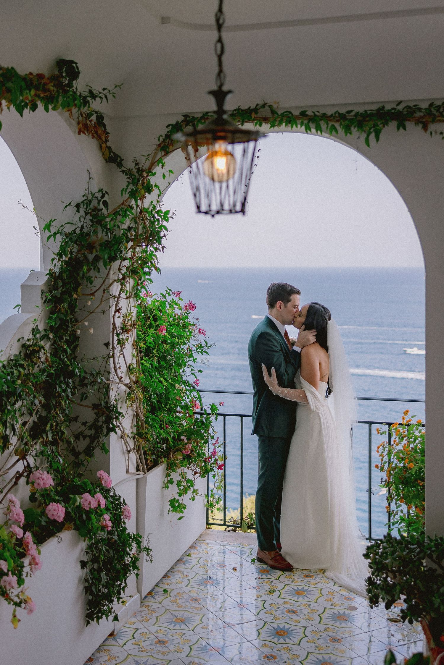 Romantic Mediterranean terrace with white archway, climbing vines, and ocean view framed by lantern and pink flowers.