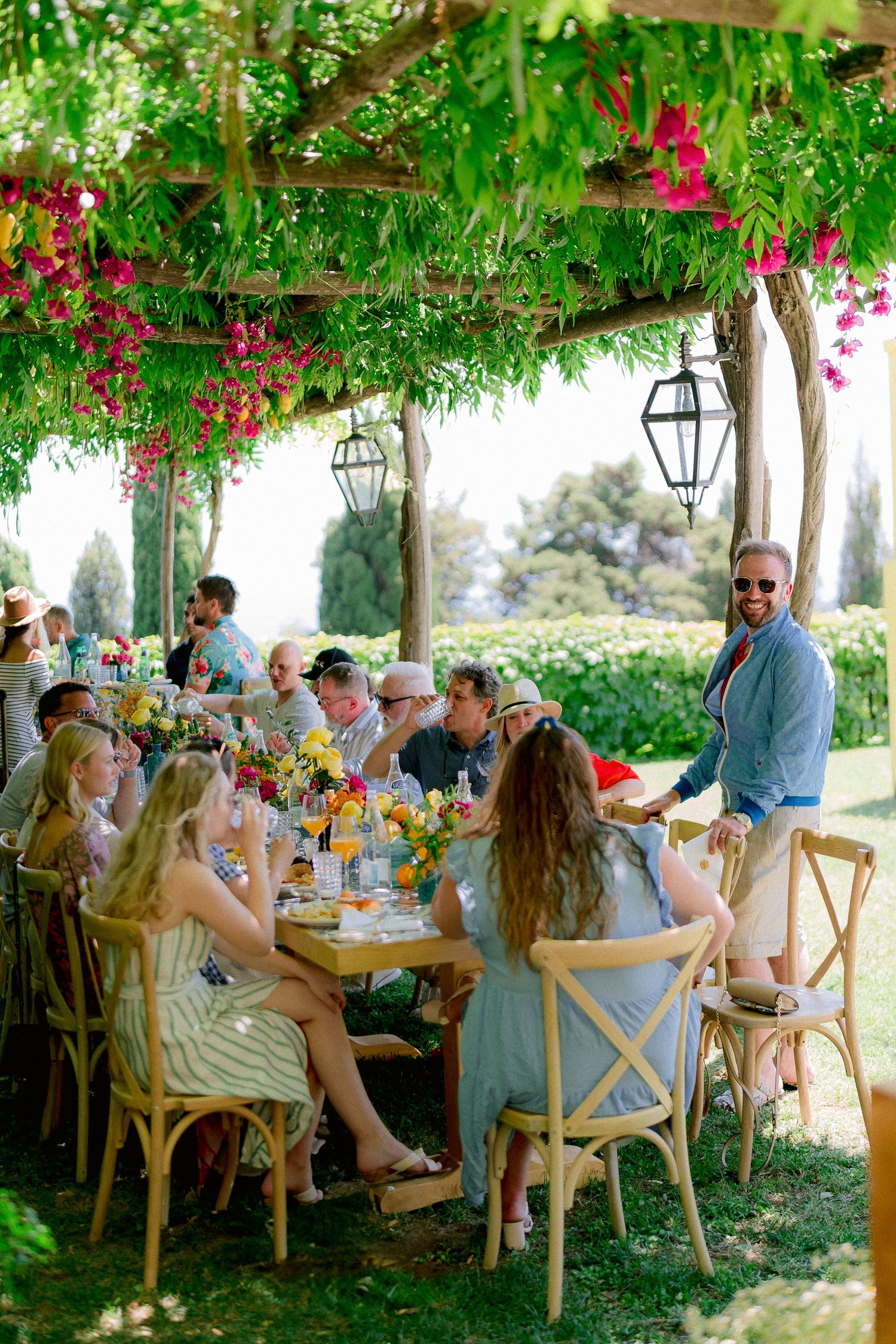 Guests enjoying an outdoor lunch under a pergola adorned with hanging flowers and lanterns at an elegant garden party.