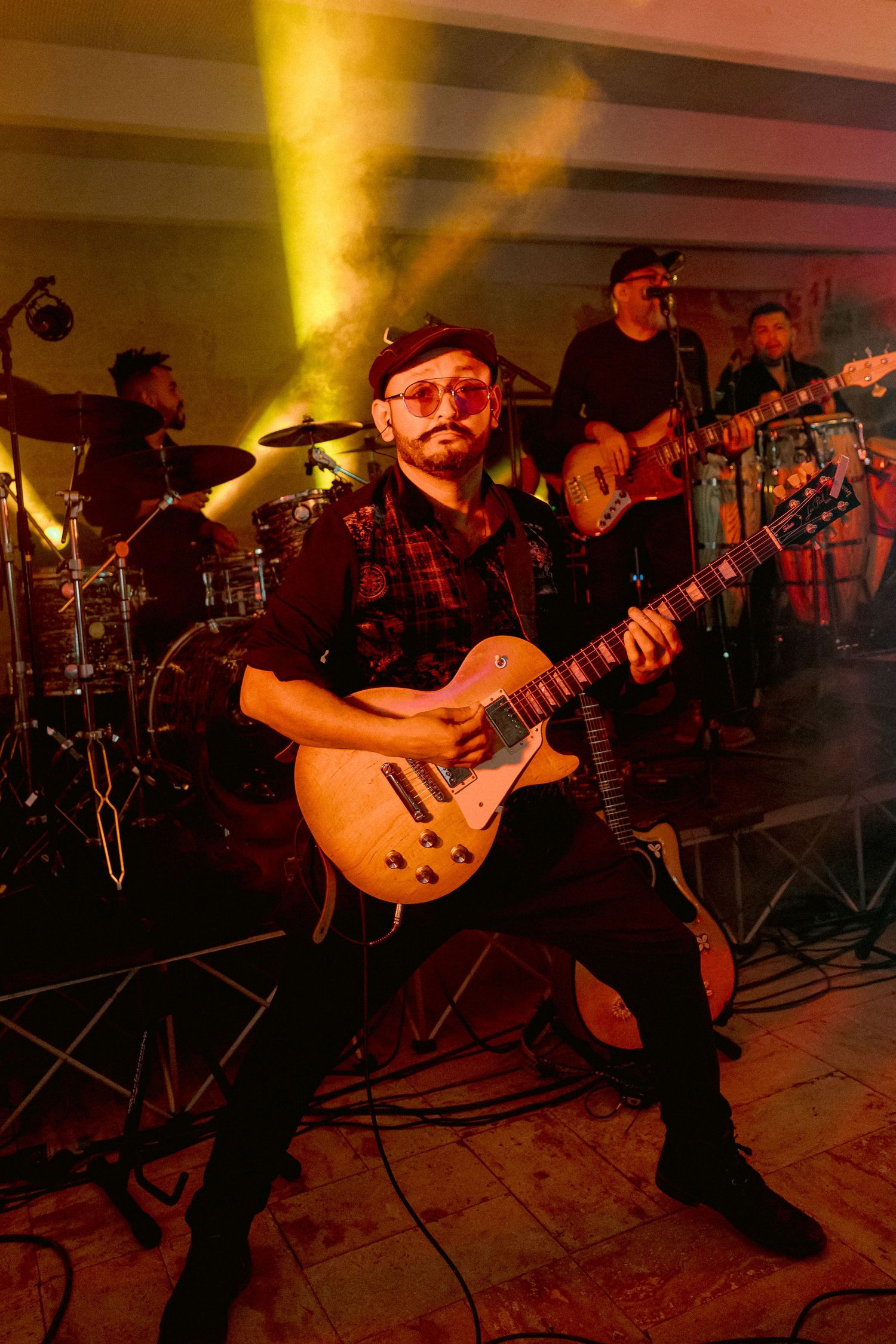 Rock band performs on stage with guitarist in foreground as yellow stage lights beam through atmospheric smoke.