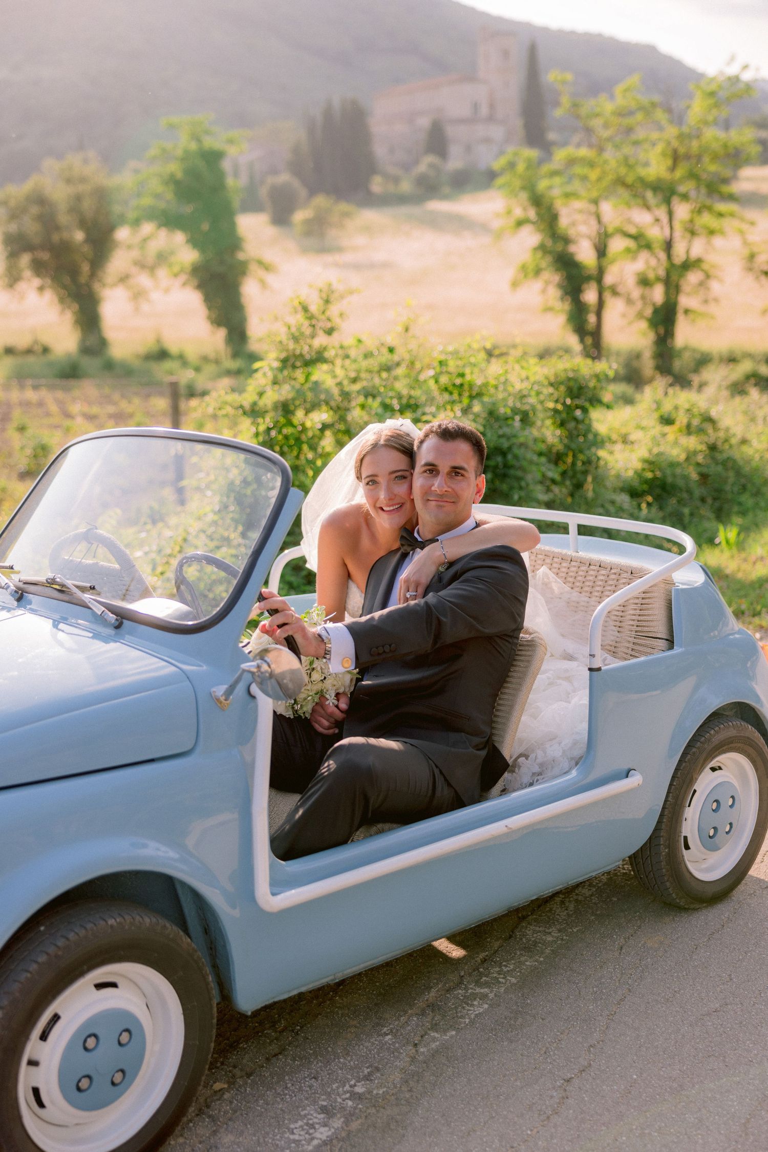 A couple sits together in a light blue vintage Fiat 500 on a scenic countryside road in summer.