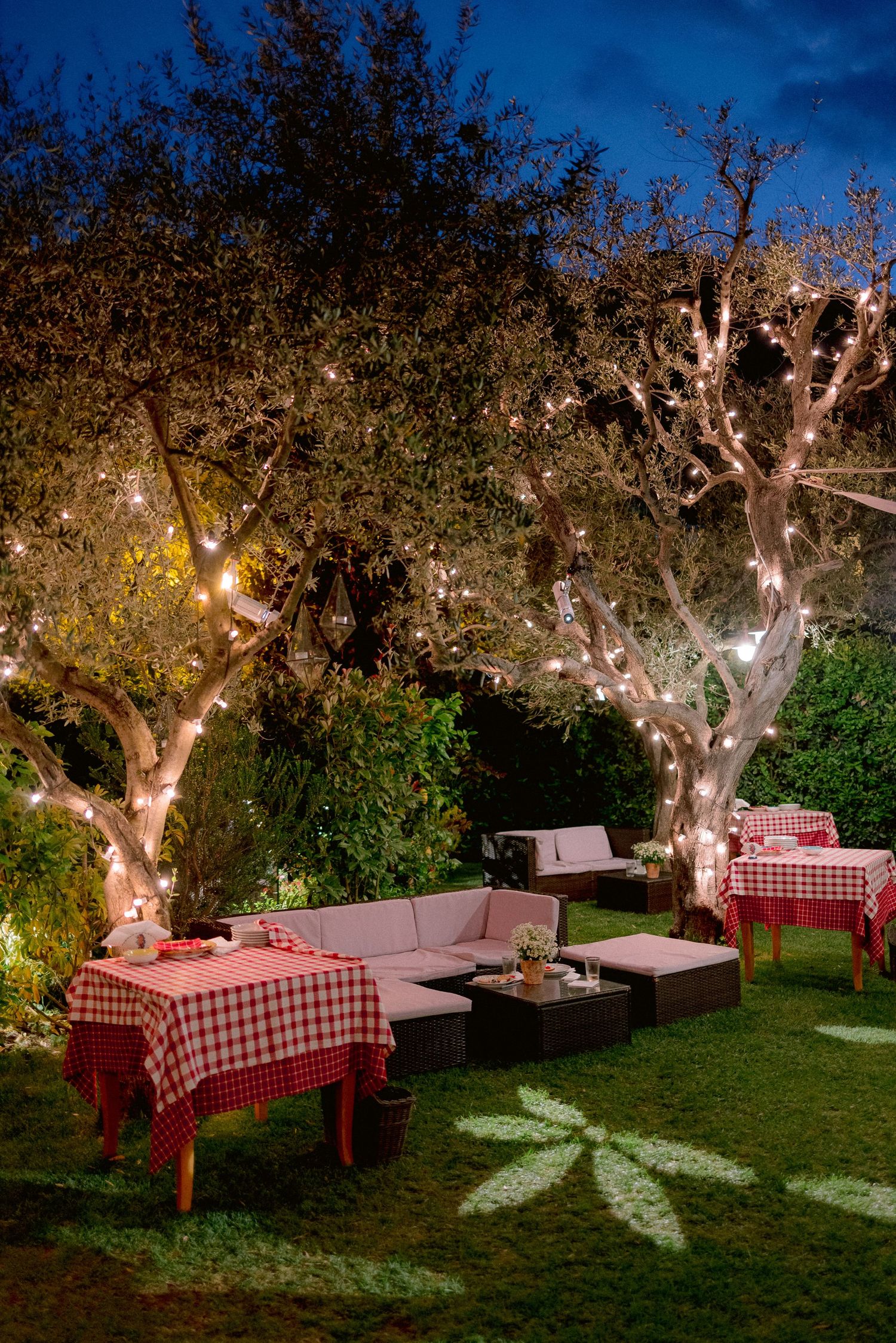 Outdoor dining setup with red checkered tablecloths and lounge seating under illuminated trees in a garden at dusk.