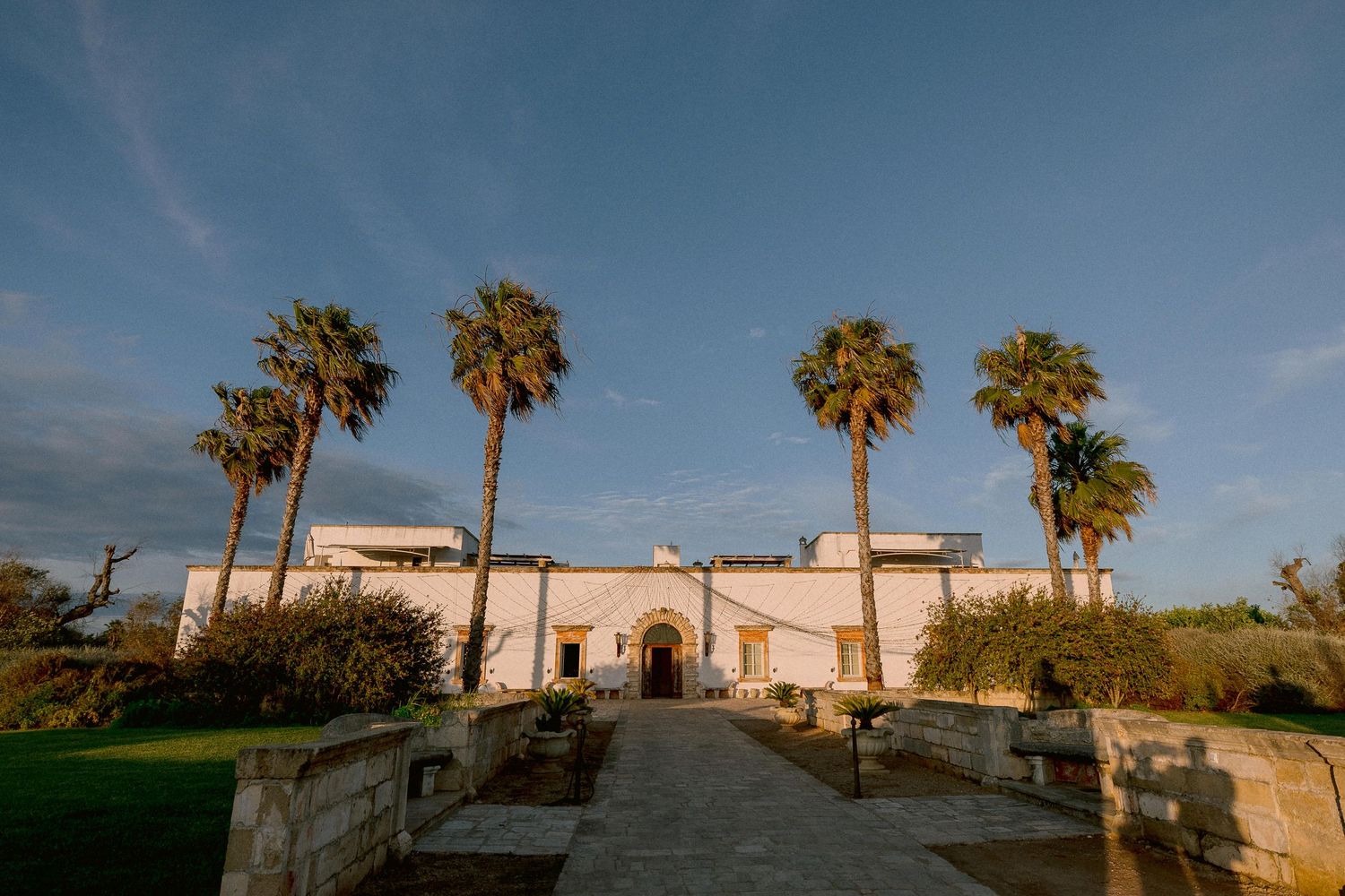 Spanish colonial style Tenuta San Mose in Salento with white walls and palm trees along a path leading to its grand entrance at sunset.