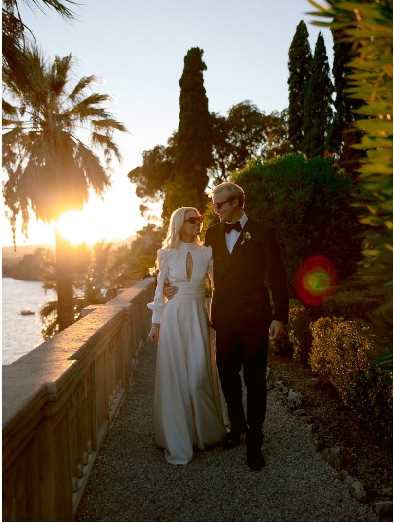 Bride in elegant white gown and groom in black tuxedo walk at sunset by waterfront garden with palm trees.