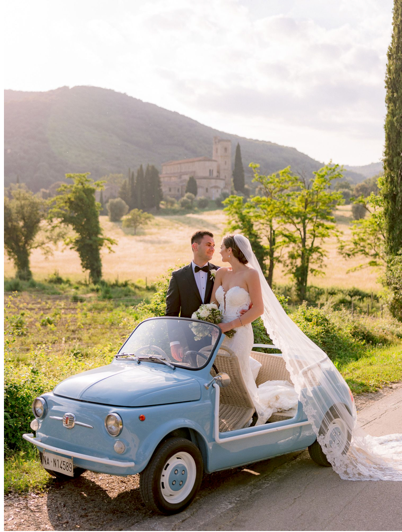 Bride and groom posing with a vintage light blue Fiat 500 in the Tuscan countryside on their wedding day.
