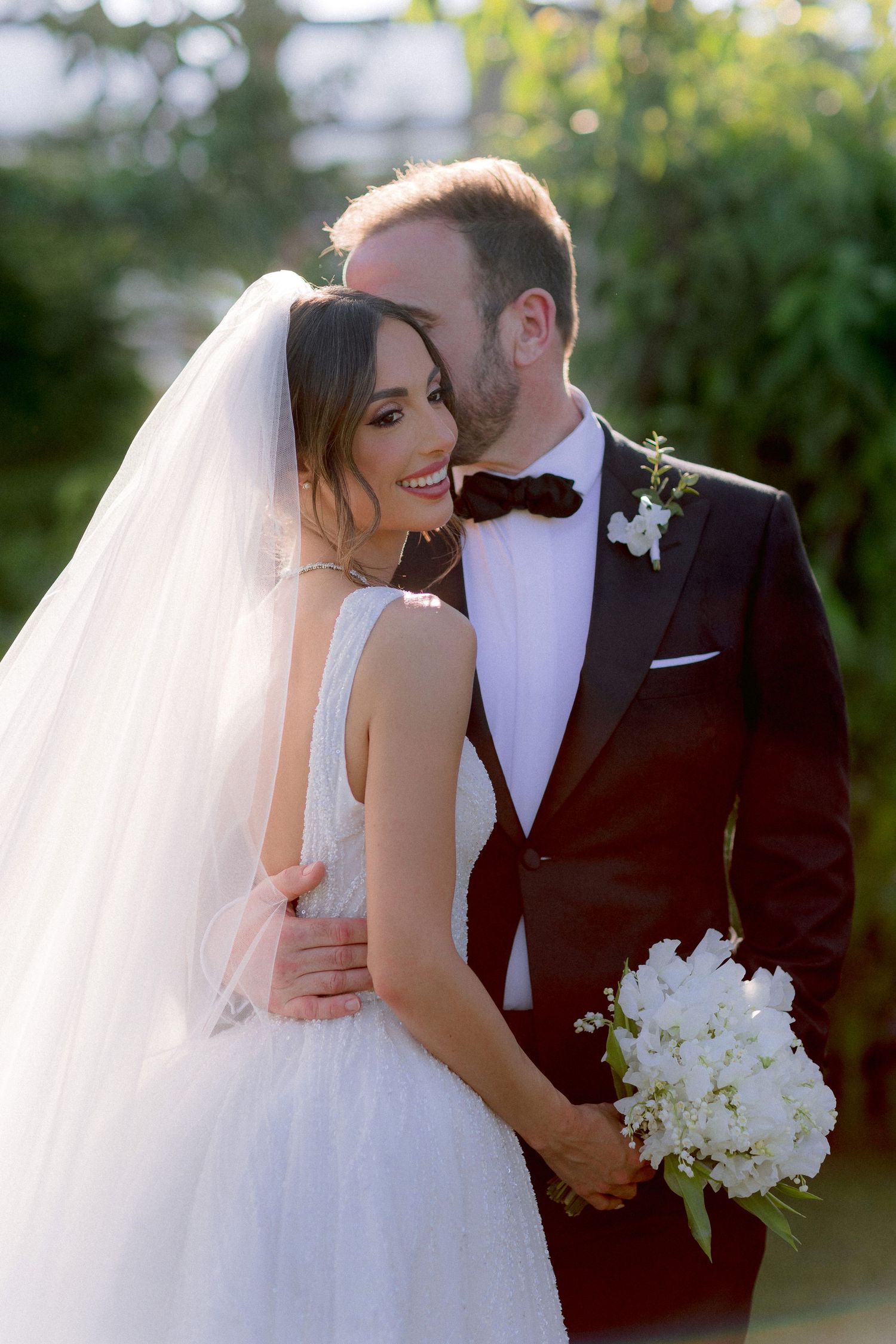 A romantic wedding portrait showing a bride in white gown with veil and groom in burgundy tuxedo embracing outdoors.