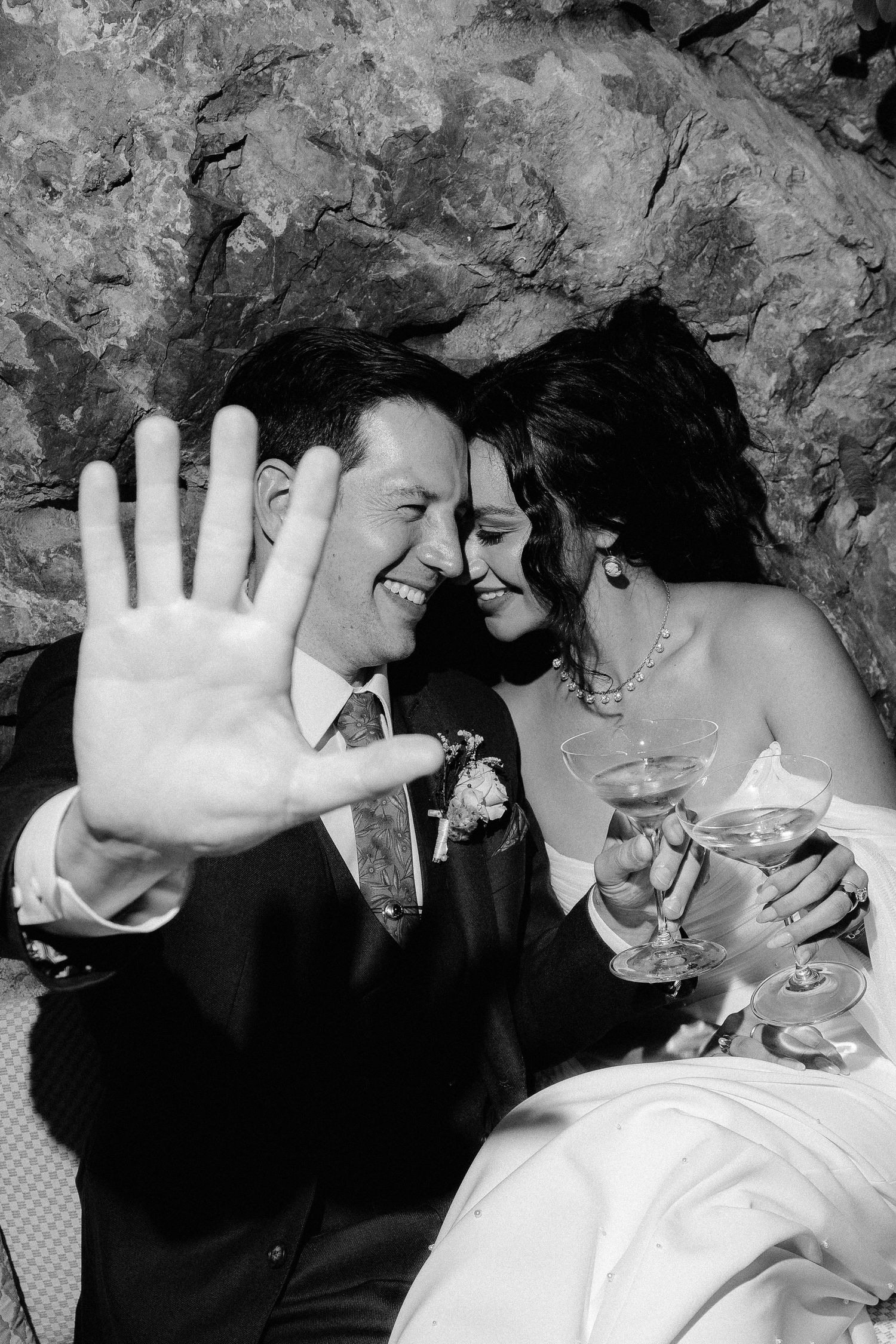 Happy couple at their wedding celebration pose playfully against a stone wall background.