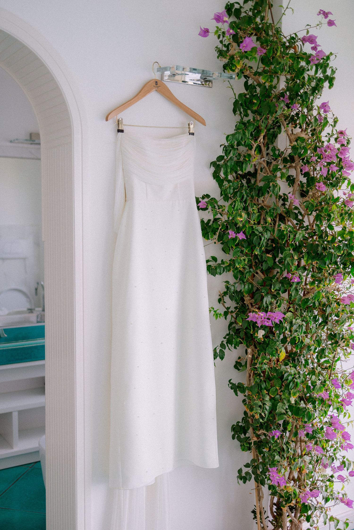 A white wedding dress hangs on a wooden hanger beside blooming pink flowers and climbing vines against a white wall.