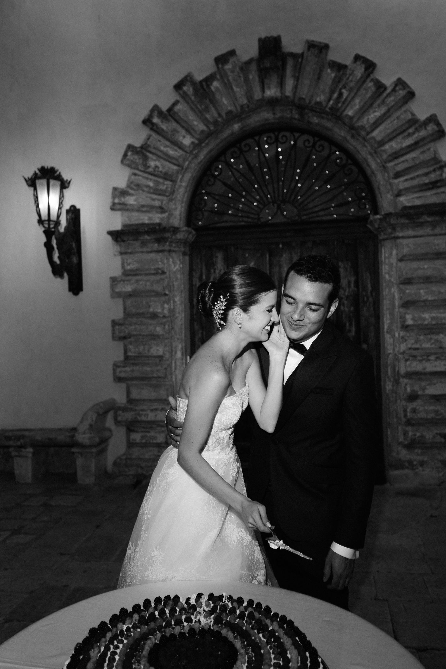 A bride and groom share an intimate moment while cutting their wedding cake under a stone archway with twinkle lights.