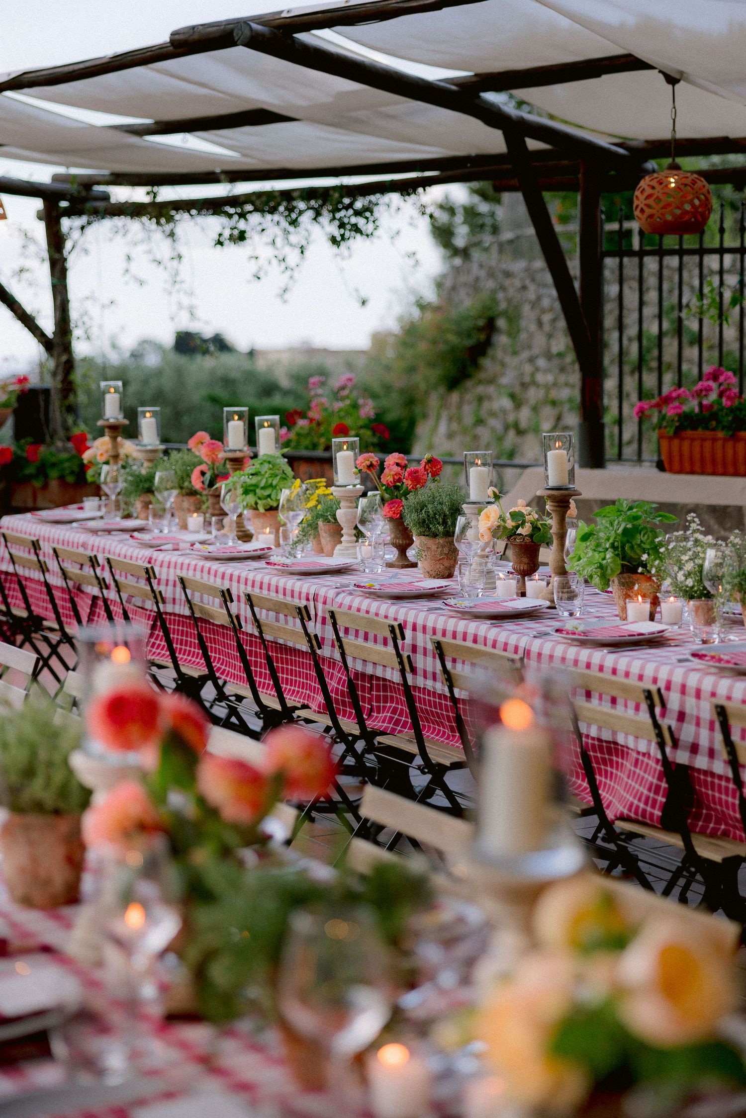 Long table with red and white checkered tablecloth set for outdoor dining with terracotta pots and candles under a pergola.