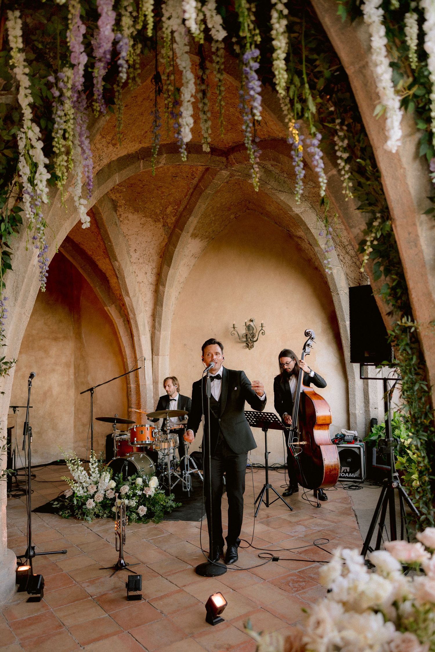 A musical band performs in an elegant arched venue with hanging wisteria flowers and decorative floral arrangements.