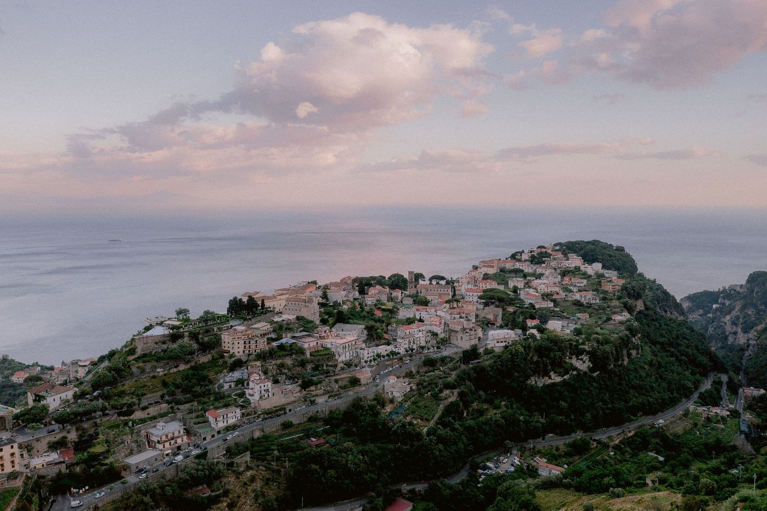 The Mediterranean coastal village of Ravello perched on mountainside overlooking calm sea at sunset with pink-tinged clouds above.